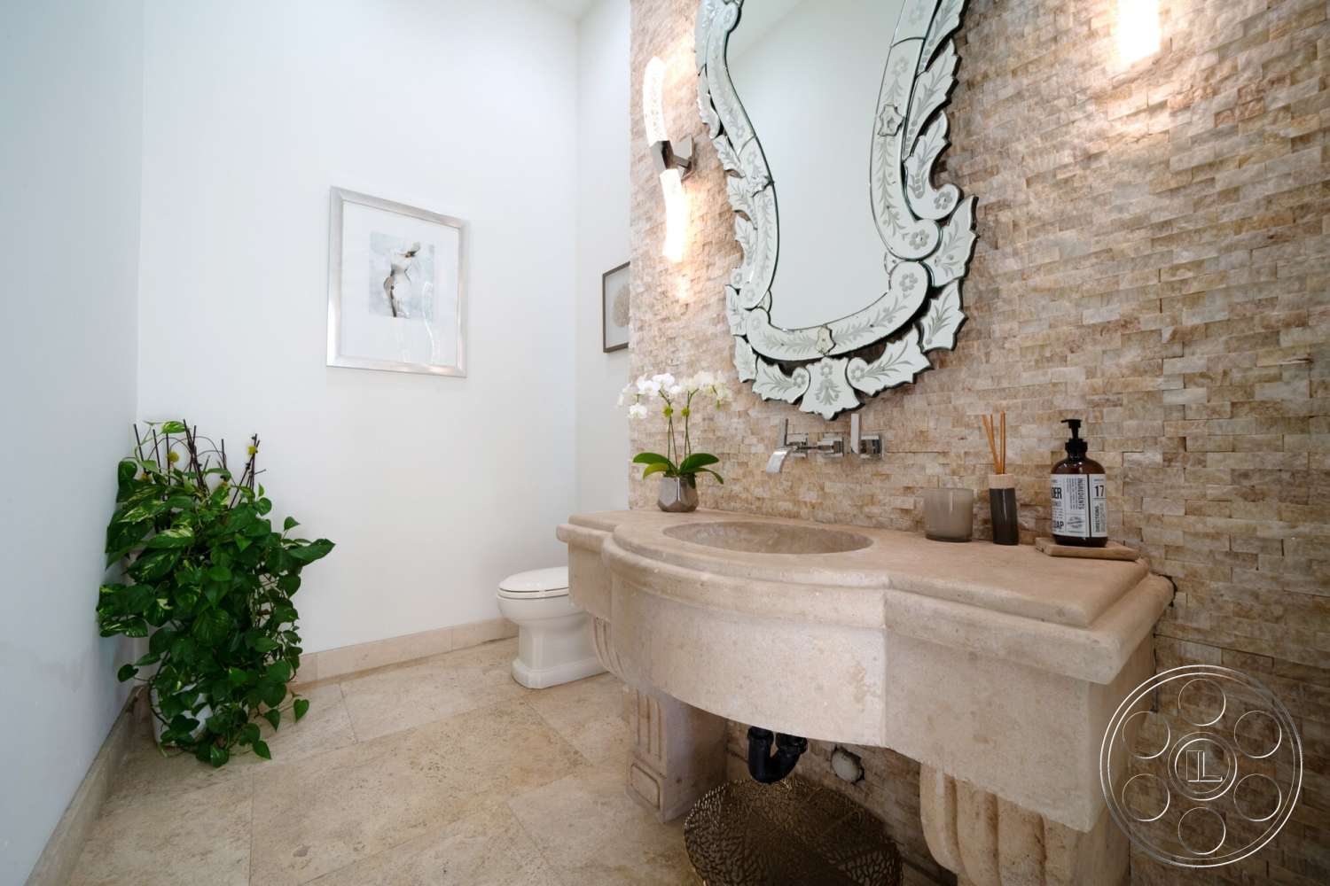Medit 169 - green potted plant, under-mounted sink, neutral painted walls, modern faucet fixtures, textured stone accent wall, light beige tile flooring, ornate wall mirror, light beige marble vanity top, single toilet