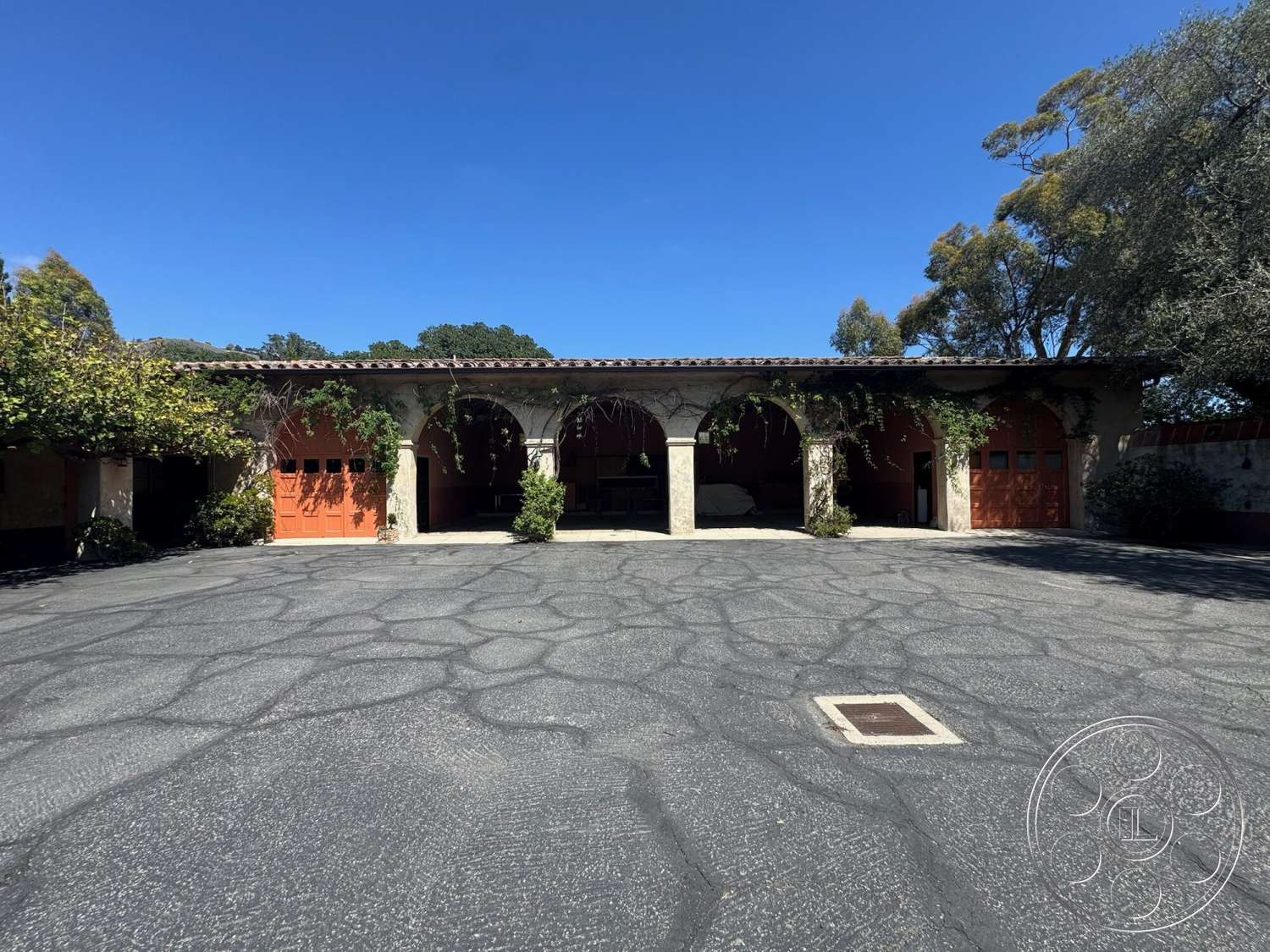 Farmstead Garden - exterior, greenery accents, arched doorways, tile roof, paved courtyard, open parking area, stone support columns, textured asphalt driveway, red wooden garage doors, outdoor vines