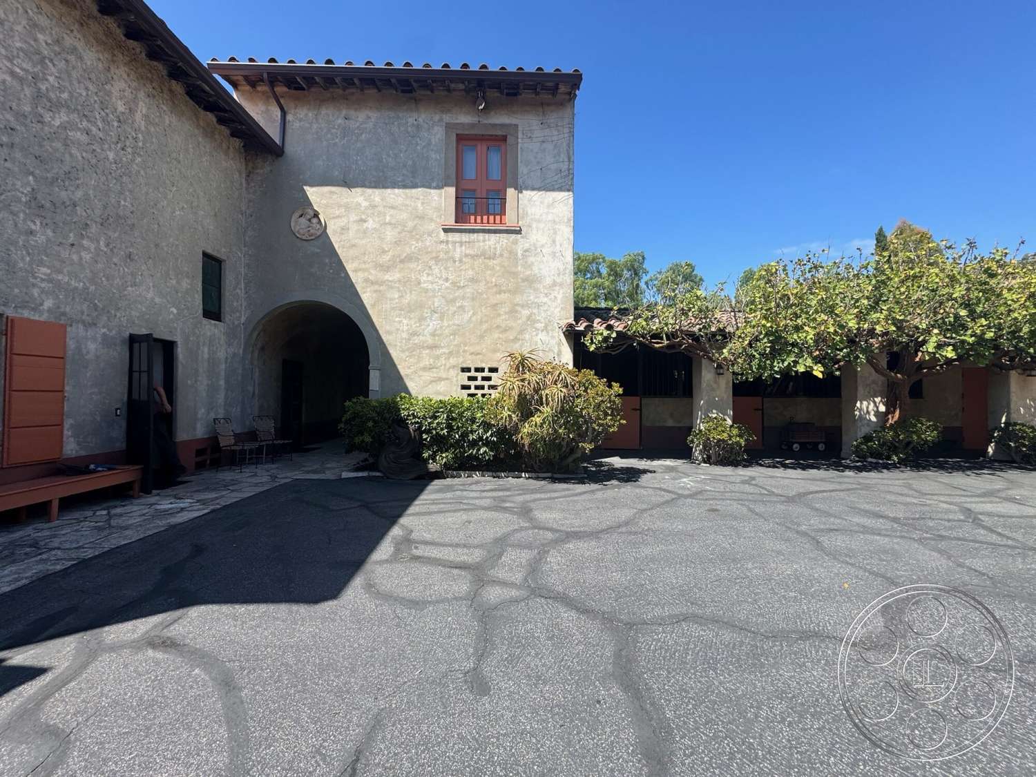 Farmstead Garden - exterior, landscaped garden, stucco walls, dark wood doors, arched entryway, tile roof, wooden window frames, paved courtyard flooring, courtyard setting, protruding tree canopy, rustic benches, red accent doors, vaulted building structure, Spanish architectural style