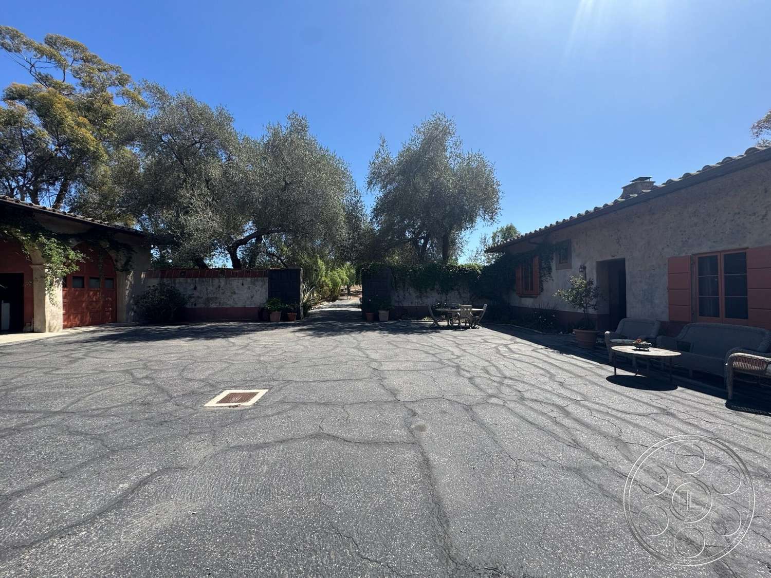 Farmstead Garden - exterior, stone wall, potted plants, outdoor seating area, landscaped courtyard, asphalt driveway, terracotta roof tiles, olive trees, whitewashed walls, Mediterranean architectural style, wooden window shutters, concrete patio area, sunlit courtyard, rustic building facade, red garage doors