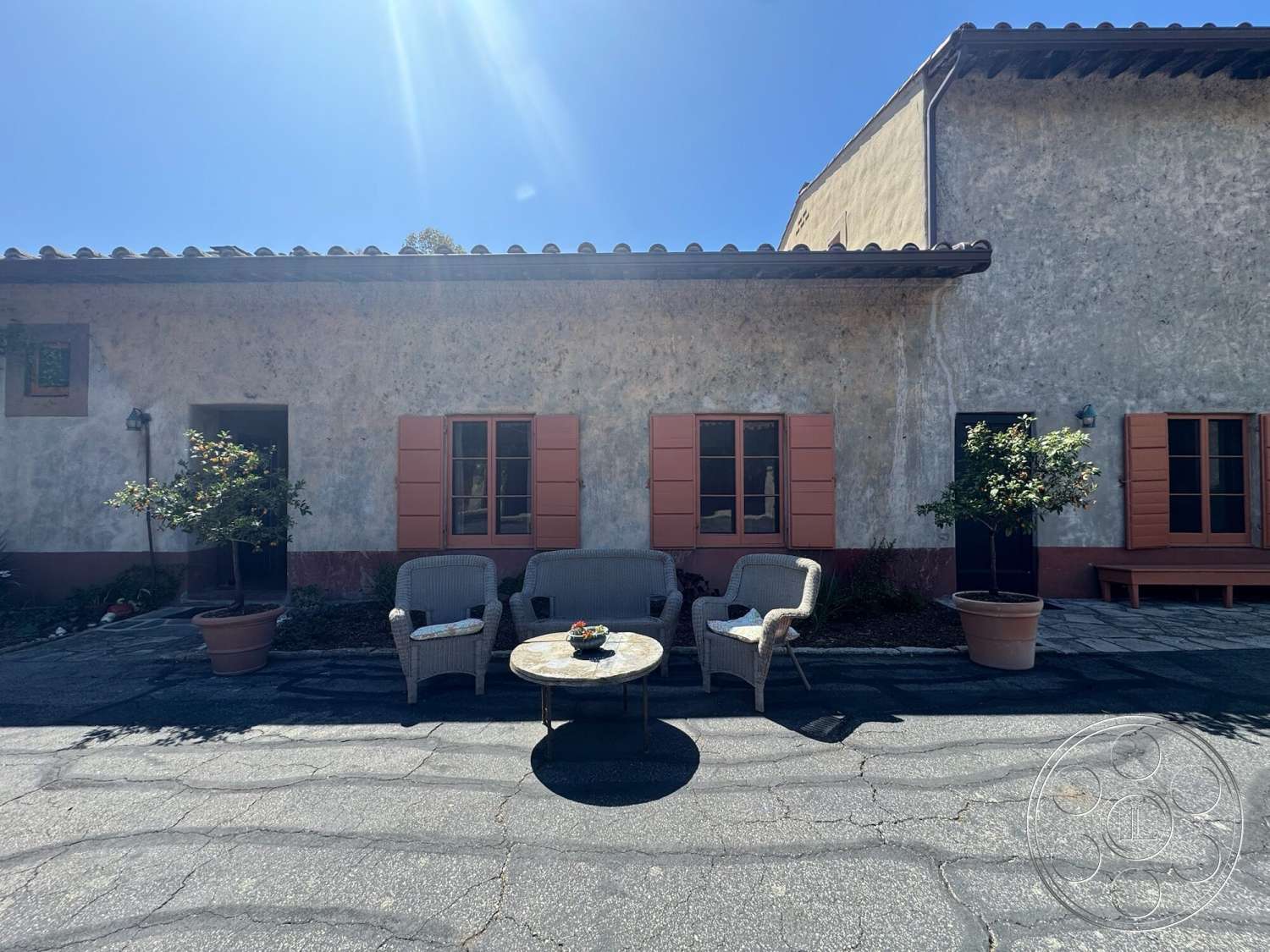Farmstead Garden - exterior, paved driveway, arched doorway, terra cotta roof tiles, modern light fixture, Mediterranean architectural style, round outdoor table, concrete patio flooring, stucco exterior wall, wicker patio chairs, landscaping with shrubs, orange window shutters, planter with small tree