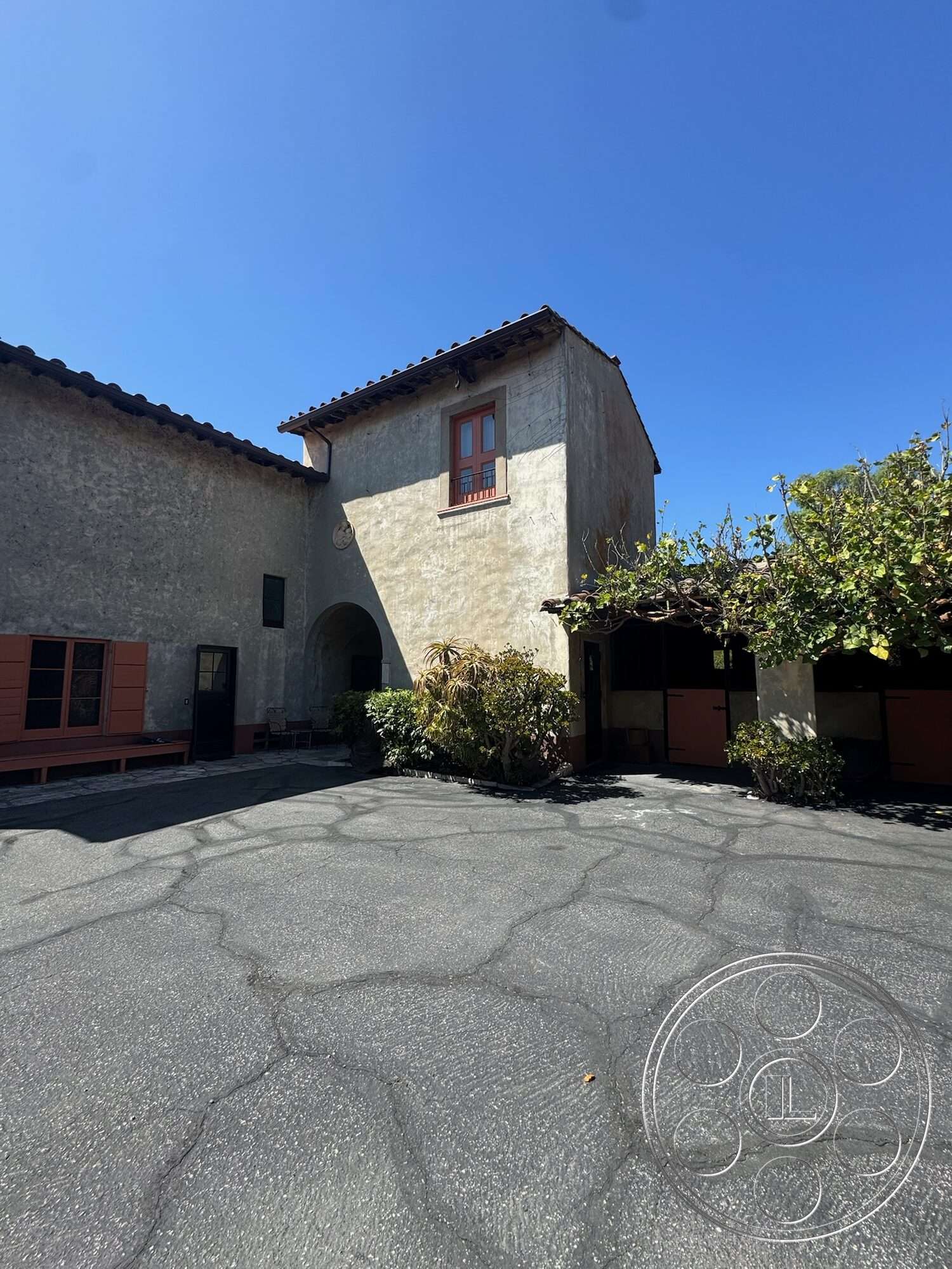 Farmstead Garden - exterior, lush greenery, arched entrance, red wooden window frames, two-story structure, landscaped garden area, stucco building facade, sloped tiled roof, paved asphalt driveway, vintage wooden doors