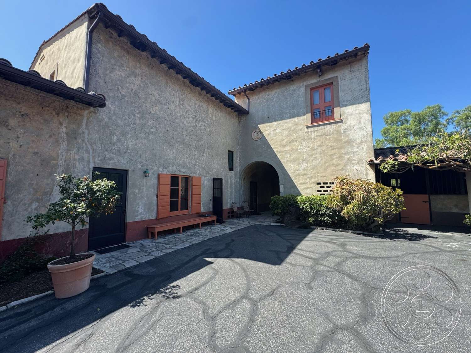 Farmstead Garden - exterior, landscaped garden, potted plant, stone pathway, concrete driveway, arched entryway, Mediterranean architectural style, clay tile roof, overhanging eaves, textured plaster wall, stucco wall finish, orange window shutters, dark wooden front door