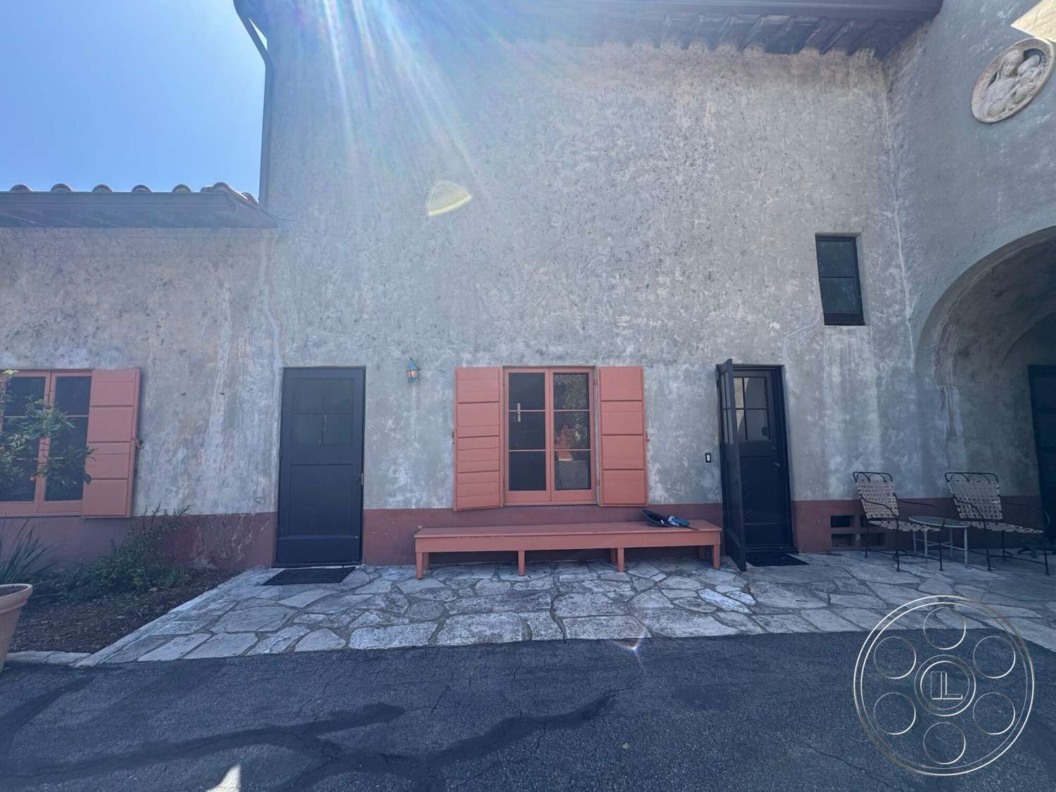 Farmstead Garden - exterior, wooden bench seating, arched doorway, landscaped garden area, stone patio flooring, plastered wall finish, orange window shutters, black paneled doors, exposed dark wood eaves, textured stucco wall treatment