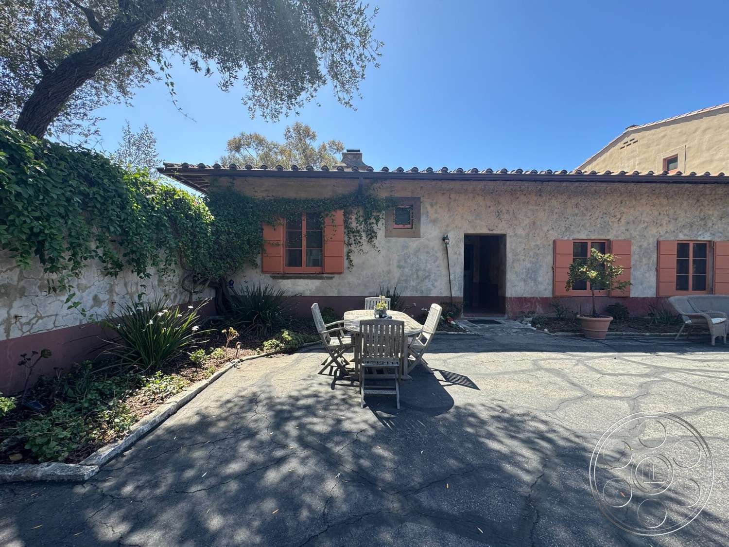 Farmstead Garden - exterior, wooden dining table, stucco walls, arched entryway, terracotta tile roof, metal outdoor chairs, landscaped garden area, flagstone patio flooring, slate stone border, orange window shutters, mature greenery, sun-drenched courtyard