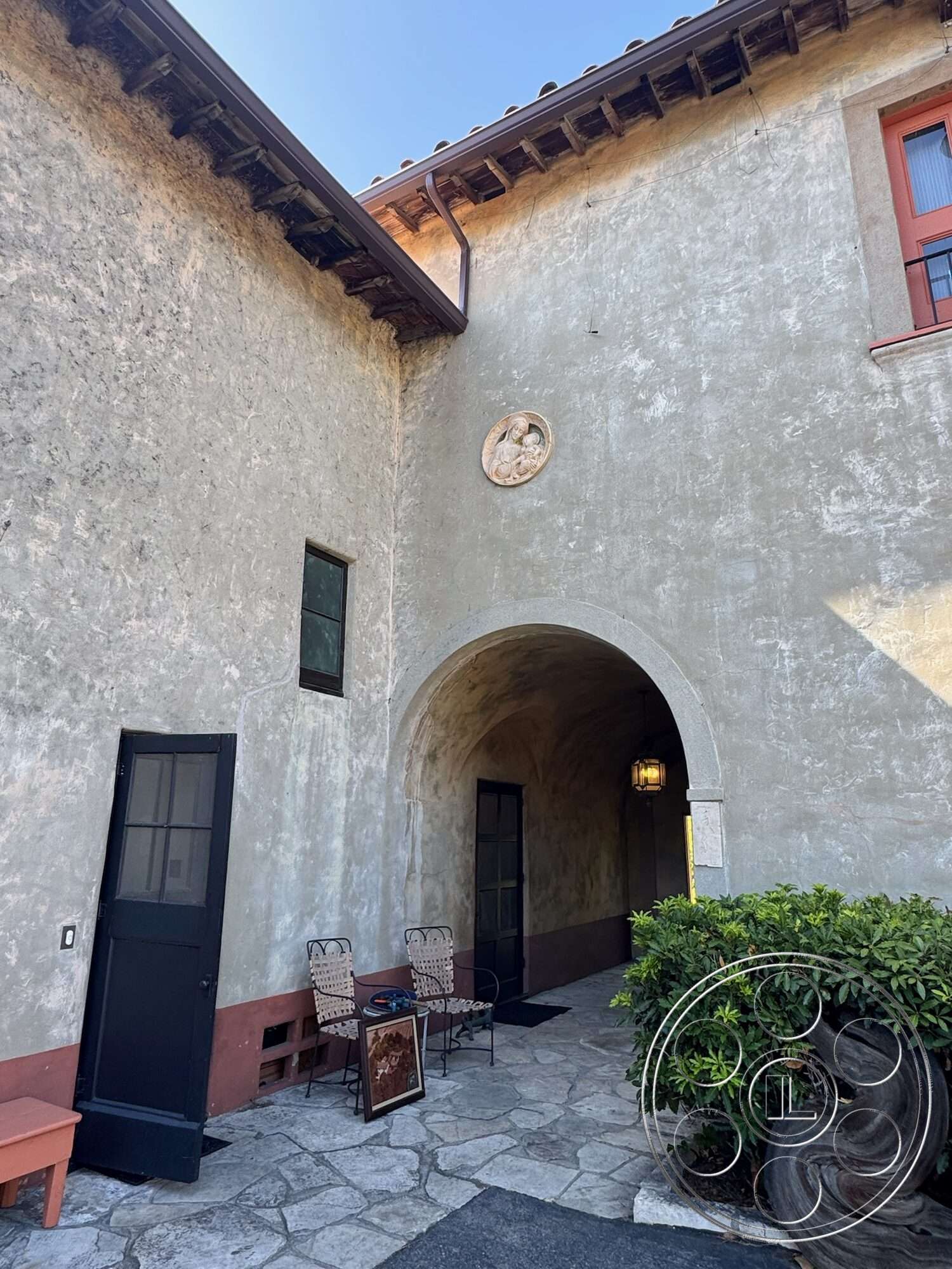 Farmstead Garden - exterior, stone pathway, traditional architecture, stucco walls, arched entryway, textured wall surface, landscaped shrubbery, courtyard setting, wooden eave details, black framed doors, window dormer, ornamental relief