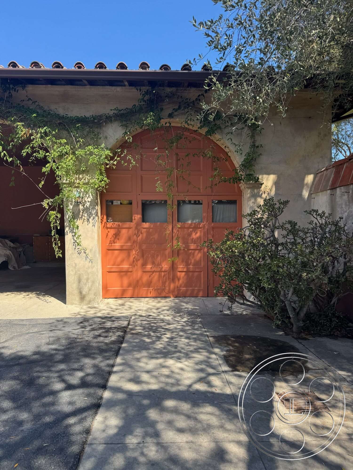 Farmstead Garden - exterior, concrete driveway, tile roof, textured plaster wall, stucco wall finish, landscaped shrubs, arched garage door, orange-painted wooden garage door, vines climbing the wall, shadow patterns on the ground, minimal garden setting