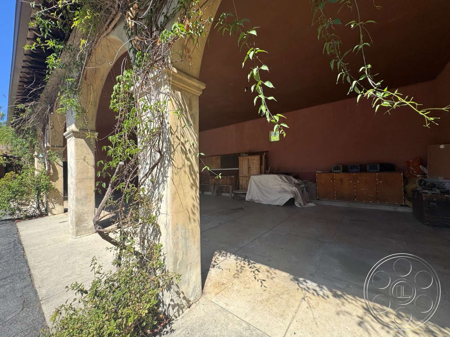 Farmstead Garden - exterior, surrounding greenery, textured plaster ceiling, arched doorways, sunlit outdoor space, exposed concrete flooring, wooden beams in ceiling, stucco walls with earthy tones, supporting stone columns, vines climbing on structural elements