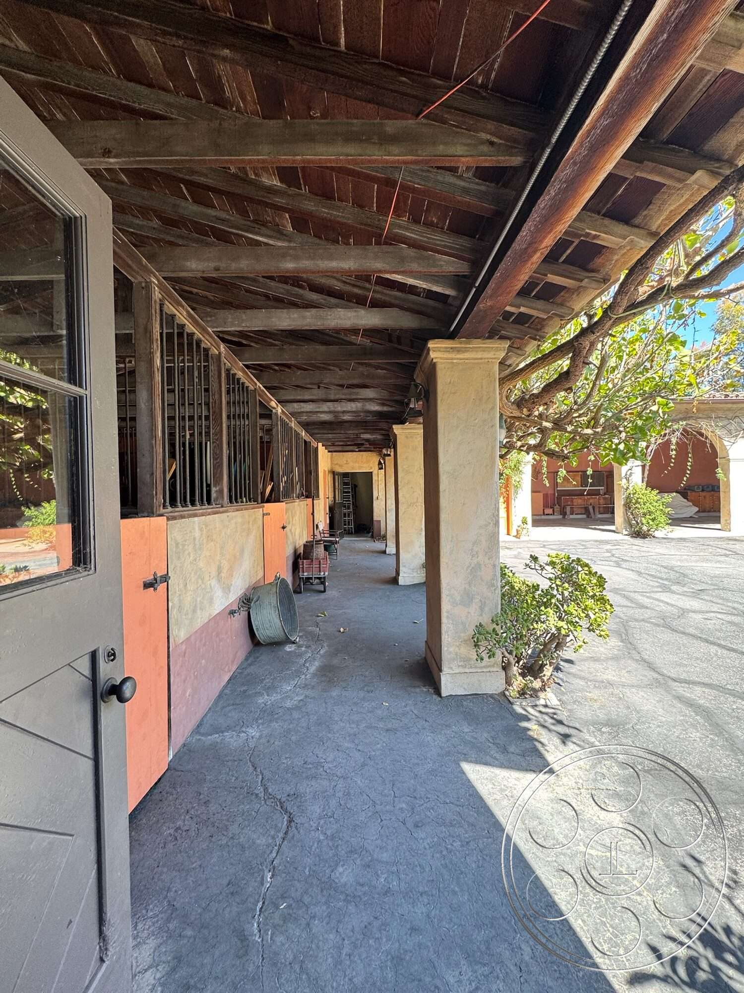 Farmstead Garden - outdoor garden area, exposed wooden beams ceiling, concrete walkway flooring, stucco wall treatments, organized horse stalls, arched passageways, dark wood door with black handle, rustic ranch-style construction, shadow play from overhead beams, plant life along walkway