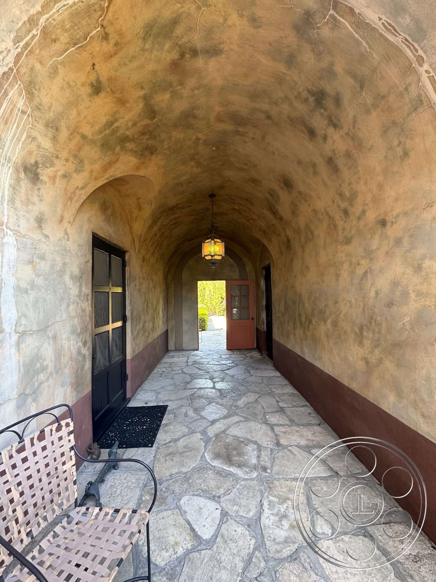 Farmstead Garden - exterior, textured plaster walls, arched entryway, hanging lantern light fixture, rustic bench seating, black metal doors, stone pathway flooring, brown stucco wall treatment