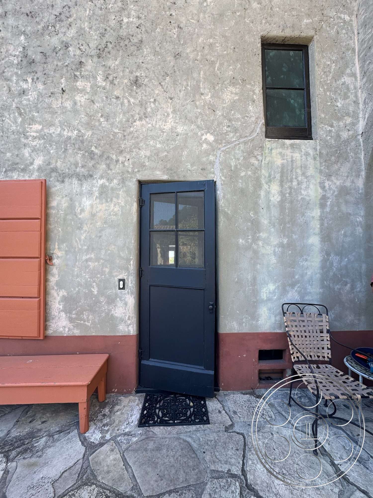 Farmstead Garden - exterior, stucco wall treatment, textured plaster finish, flagstone patio flooring, metal outdoor chair, black wooden door with glass panels, small rectangular window, orange painted wall panel, brown painted base, decorative door mat
