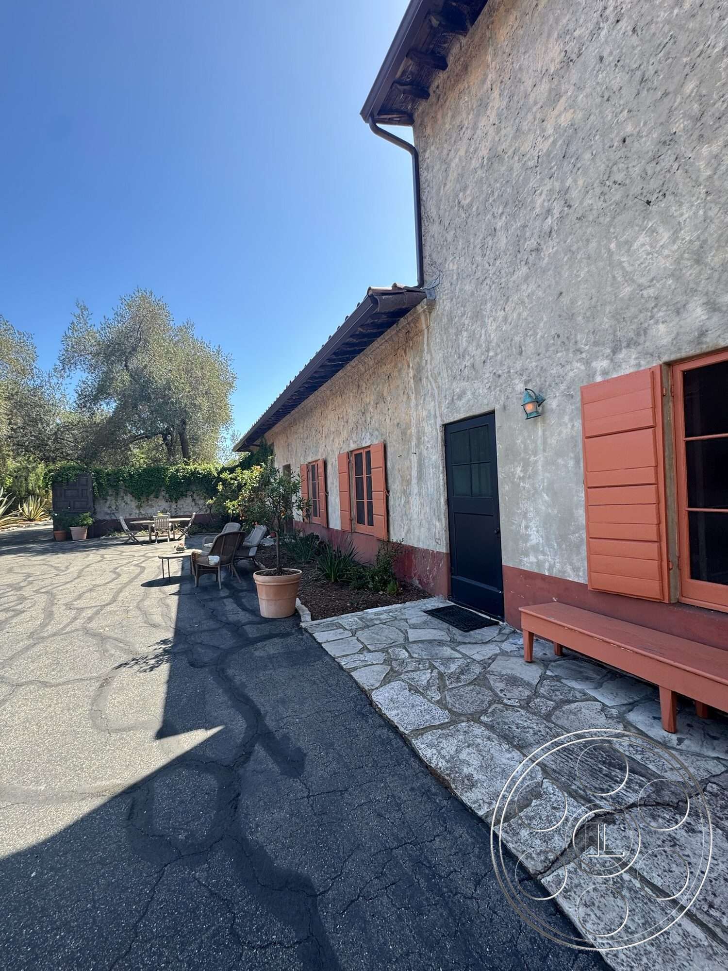 Farmstead Garden - exterior, landscaped garden, wooden bench, outdoor seating area, asphalt driveway, stucco walls, dark wood door, vintage light fixture, rustic architectural style, stone patio flooring, courtyard setting, orange wooden shutters