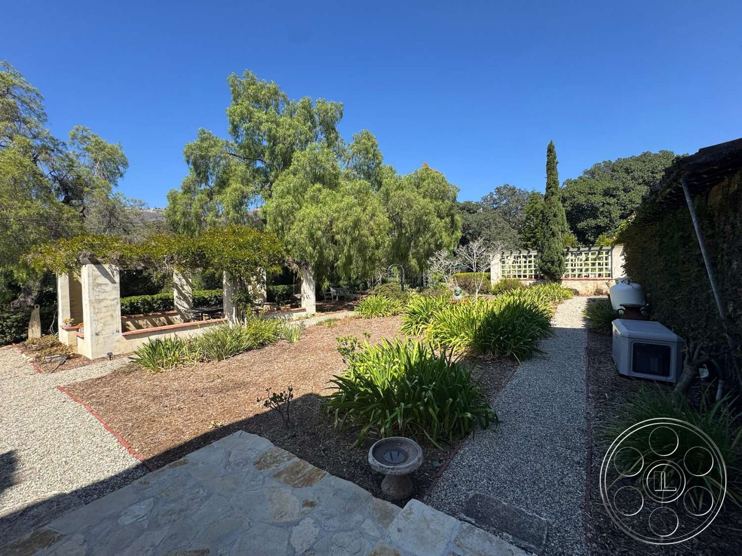 Farmstead Garden - exterior, outdoor seating area, gravel pathway, lush vegetation, wooden pergola, natural stone features, sunny outdoor setting, textured garden beds, stone patio flooring, softscape garden design, native landscaping, greens and bushes, vertical wooden trellis