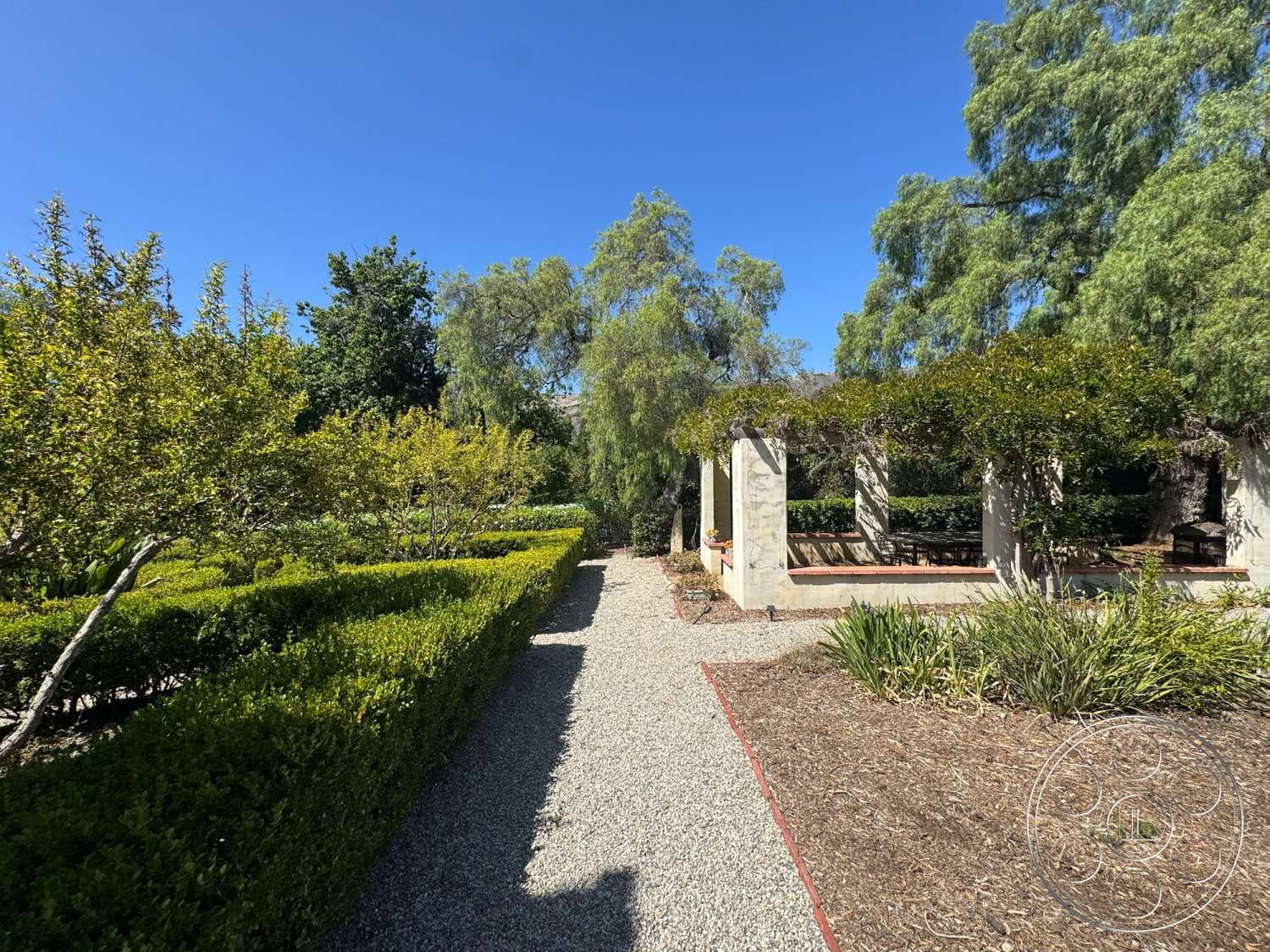 Farmstead Garden - exterior, gravel pathway, landscape plants, boxwood hedges, wooden benches, lush green foliage, open sky view, raised flower beds, pergola with concrete columns, textured earth mulch