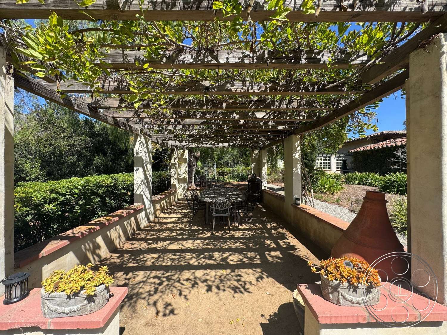 Farmstead Garden - exterior, black metal chairs, brick border, stucco walls, lush green hedges, gravel landscape, small potted plants, wood pergola, sun-drenched patio area, metal outdoor dining table, terracotta garden pot, vine-covered pergola