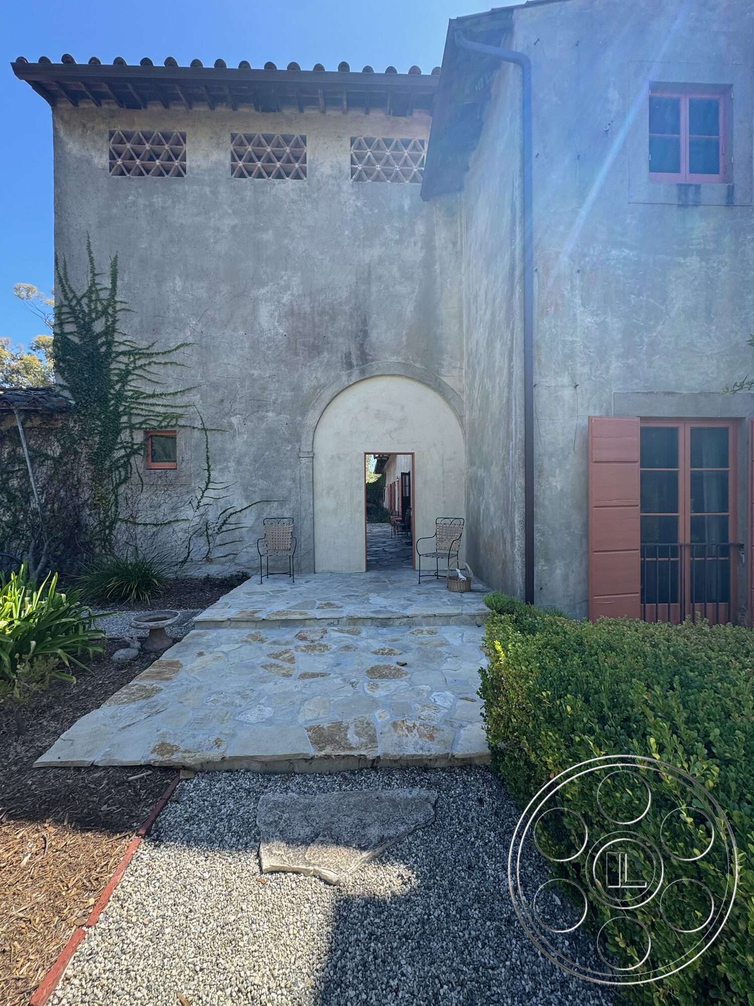 Farmstead Garden - exterior, outdoor seating area, stucco walls, lush green hedges, arched entrance, gravel landscape, mediterranean architectural style, slate stone walkway, brick window details, wood eave overhang, red wooden windows