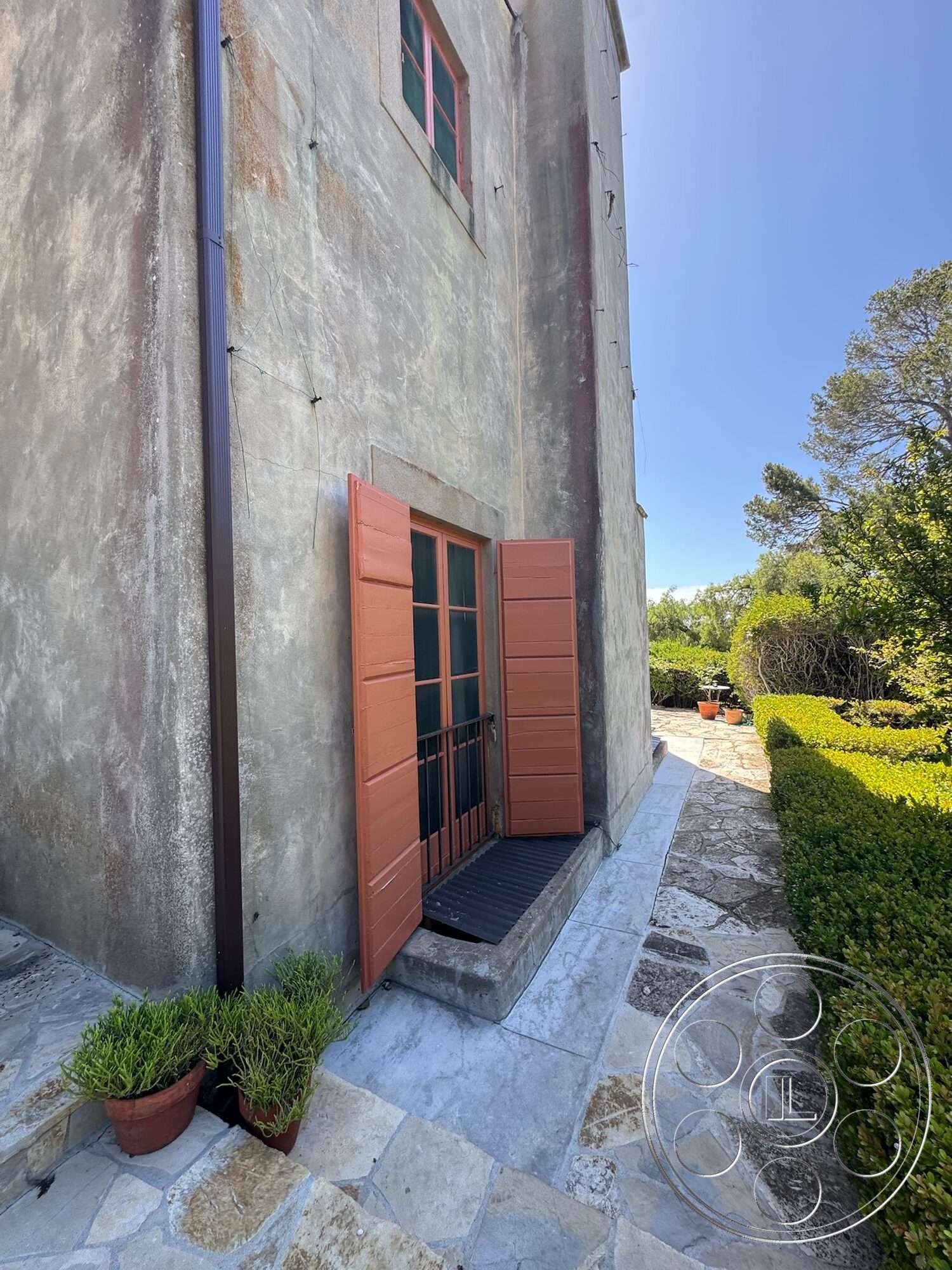 Farmstead Garden - exterior, landscaped garden, potted plants, stone pathway, stucco walls, metal downspout, green hedge border, double hung windows, circular stone steps, orange wooden shutters, concrete window sill