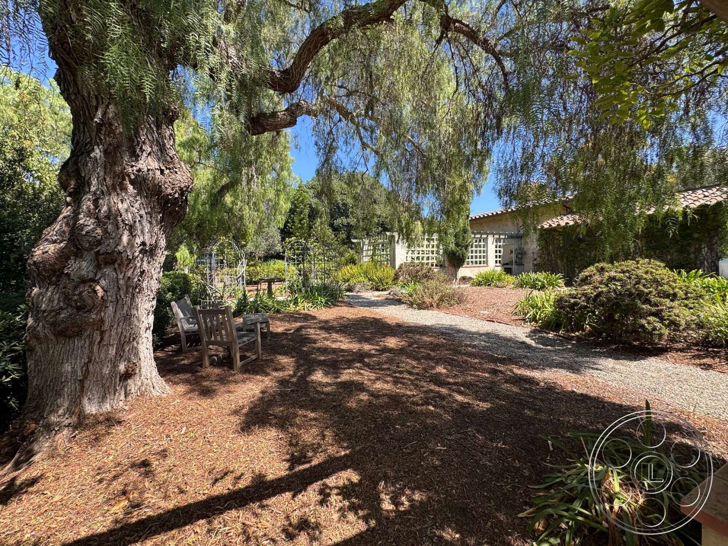 Farmstead Garden - exterior, landscaped garden, gravel pathway, garden setting, wooden garden bench, paved patio area, stucco exterior wall, textured ground cover, dappled sunlight through leaves, mature tree with textured bark, lattice trellis, greenery including shrubs and plants, tile roof details