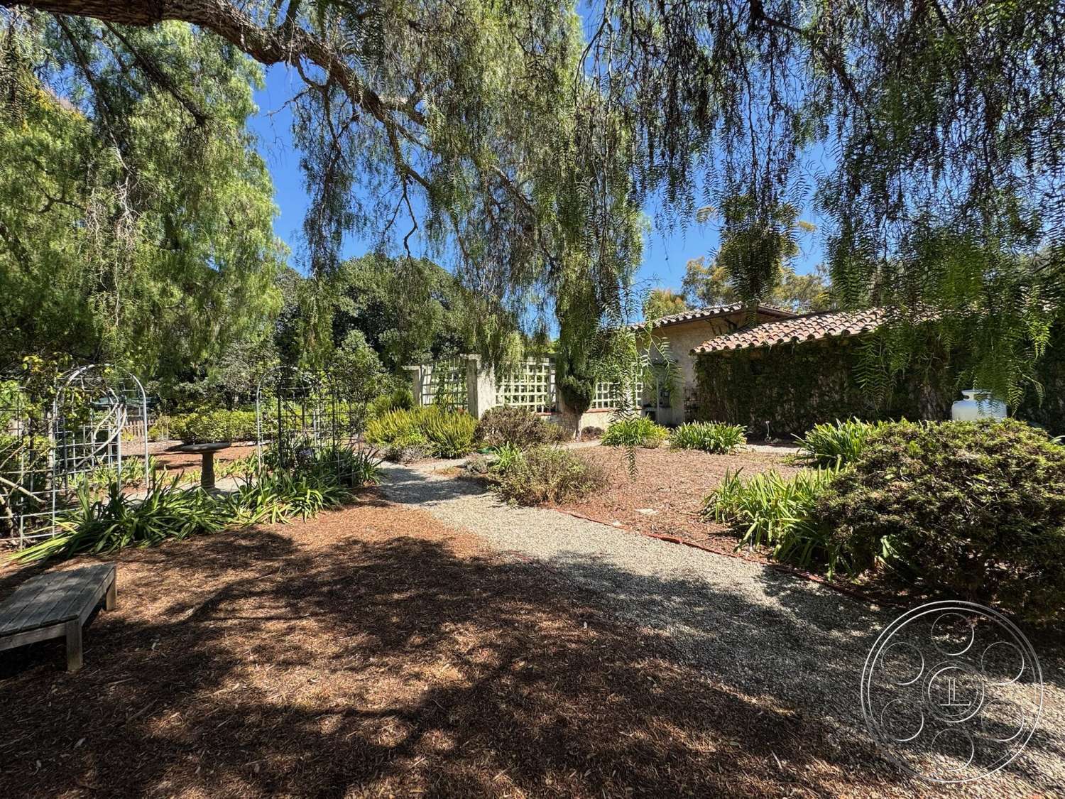 Farmstead Garden - exterior, stone wall, lush landscaping, gravel pathway, Mediterranean architecture, decorative garden elements, dappled sunlight through trees, pergola structure, wood garden bench, woven trellis, plant beds with diverse foliage, tile roof on house, airy outdoor space, rustic wooden elements, natural mulch ground cover