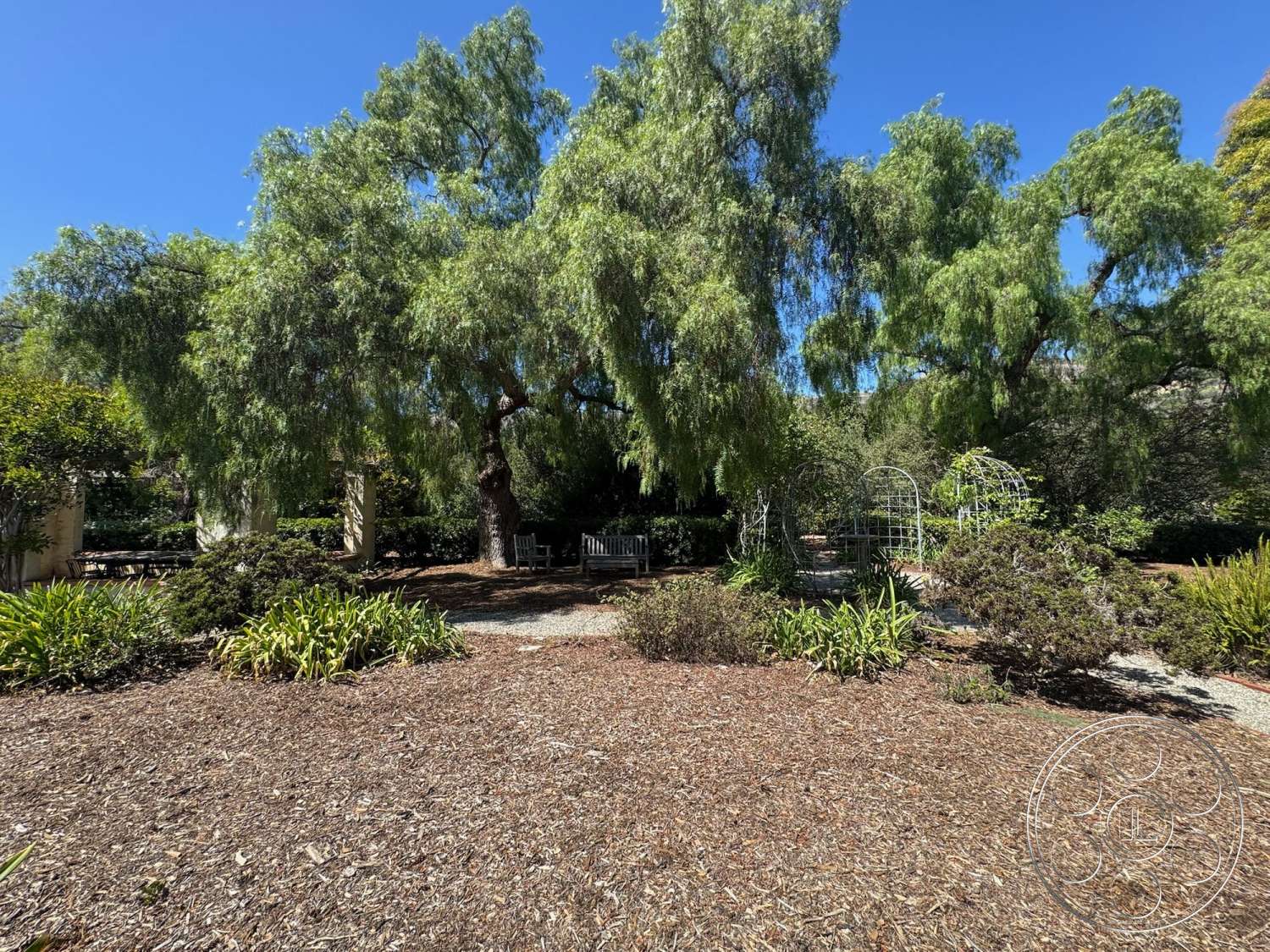 Farmstead Garden - exterior, lush greenery, outdoor seating area, gravel pathway, wooden benches, landscaped garden beds, sunlit garden setting, mature shrubbery, pergola structure, large leafy trees, metal garden trellises, mulched ground cover