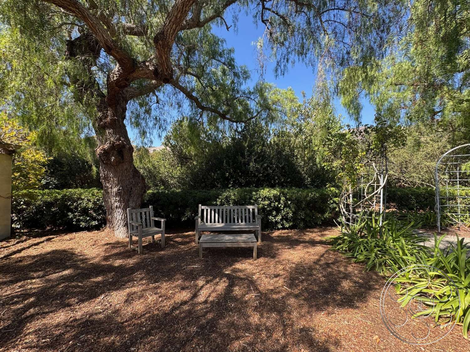 Farmstead Garden - exterior, landscape plants, lush green hedges, sunlit outdoor space, natural mulch ground cover, wooden garden benches, mature shade tree canopy, metal trellis structure, aromatic herb garden