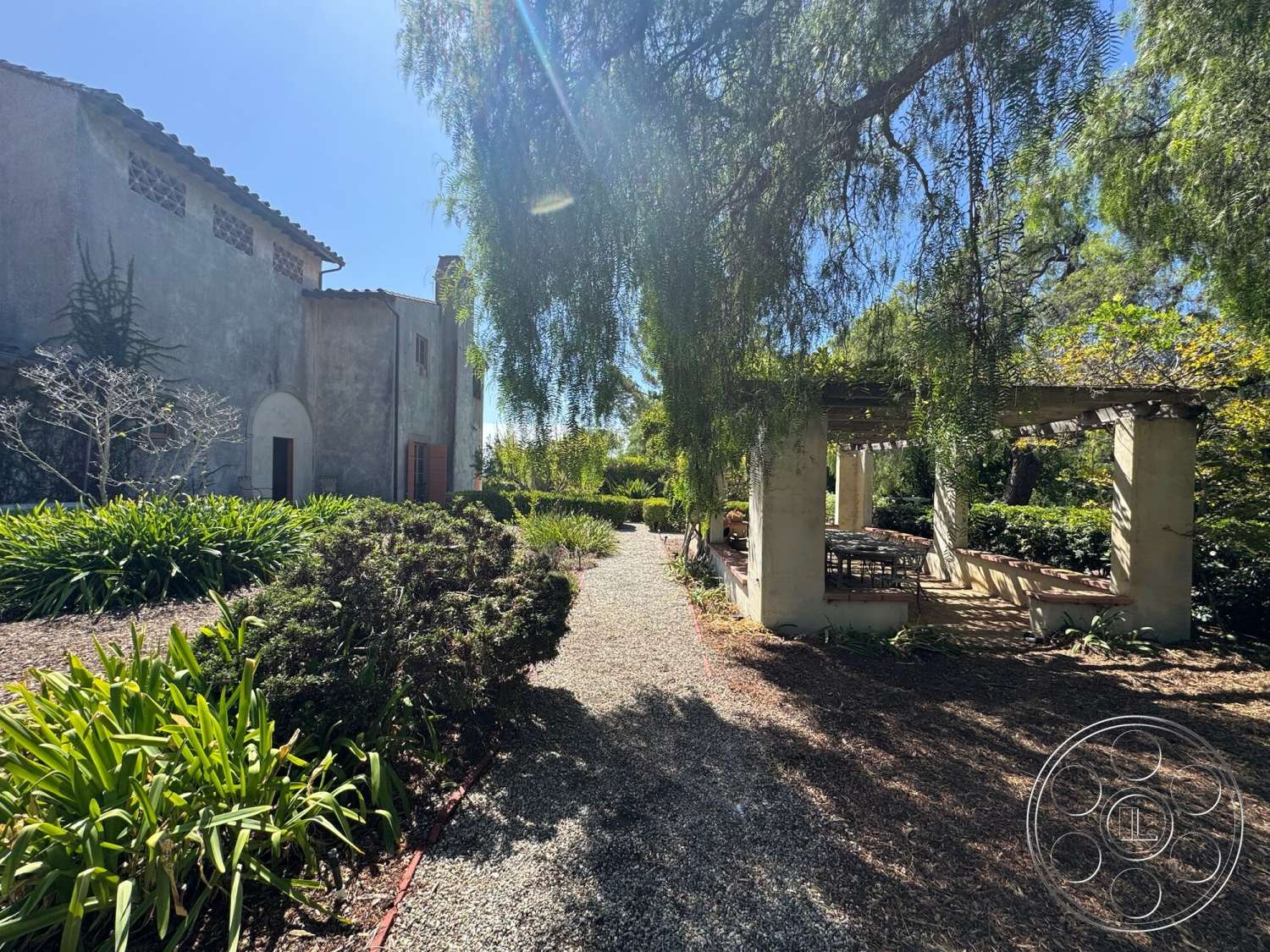 Farmstead Garden - exterior, lush greenery, gravel pathway, terracotta roof tiles, stucco walls, arched doorways, rustic outdoor dining area, landscaped garden area, pergola with concrete columns, decorative stone edging, painted wooden shutters