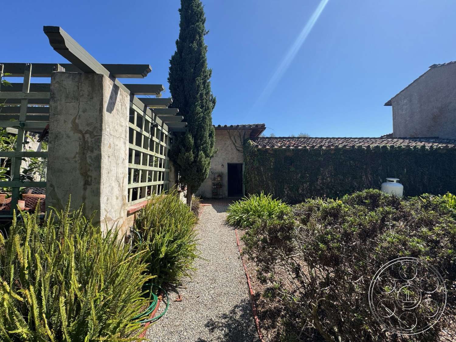 Farmstead Garden - exterior, outdoor seating area, gravel pathway, terracotta roof tiles, stucco walls, cypress trees, ivy-covered wall, decorative stone border, lush greenery landscaping, pergola with wooden beams, simple garden design