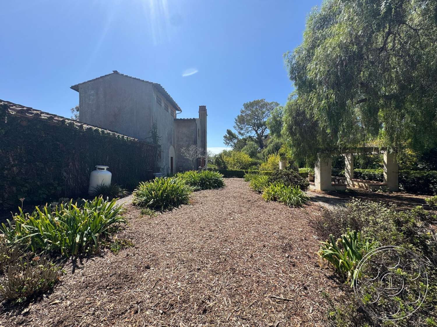Farmstead Garden - exterior, landscaped garden, lush greenery, wooden pergola, residential backyard, mulch ground cover, garden seating area, clay tile roof, stone feature wall, sunny outdoor setting, stucco house exterior, vines on fence