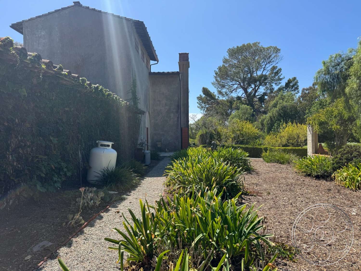 Farmstead Garden - exterior, gravel pathway, wooden planter boxes, large trees, terracotta accents, tile roof, tall hedges, stucco wall, landscape garden, vegetation border, sunlit garden setting, natural stone feature, gas tank in garden, softscaping with shrubs
