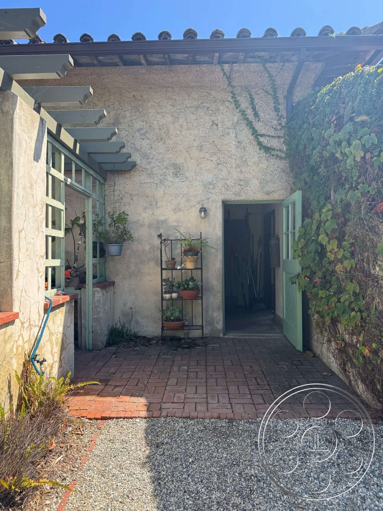 Farmstead Garden - exterior, gravel landscaping, terracotta tile roof, stucco wall finish, outdoor garden area, red brick paver pathway, pergola structure, green wooden door, potted plant arrangement, ivy plant growth