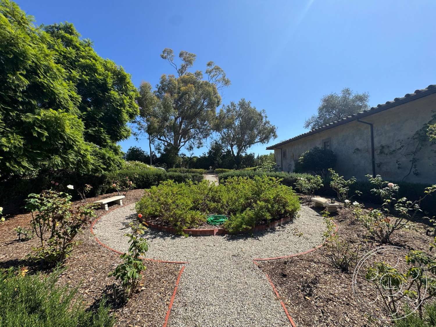 Farmstead Garden - exterior, wooden bench, tile roof, brick garden border, stucco house exterior, landscaped garden beds, mature trees providing shade, lush green shrubs, sunny courtyard setting, flowering rose bushes, rounded gravel pathway