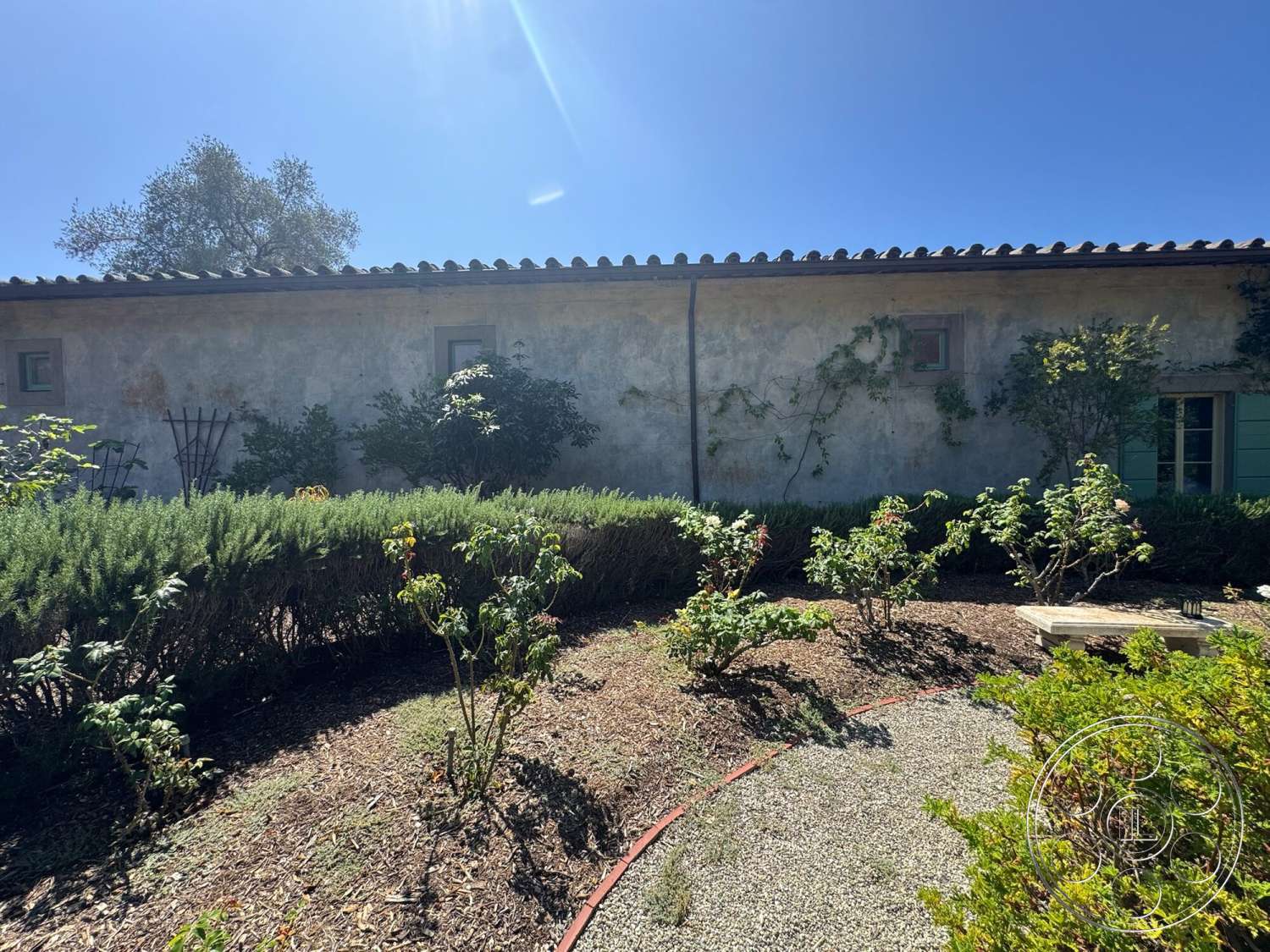 Farmstead Garden - exterior, surrounding greenery, landscape plants, terracotta roof tiles, green wooden windows, stucco wall finish, gravel garden path, rustic garden setting, blue sky backdrop, vine-covered wall, ornamental garden trellis, flower bed with shrubs