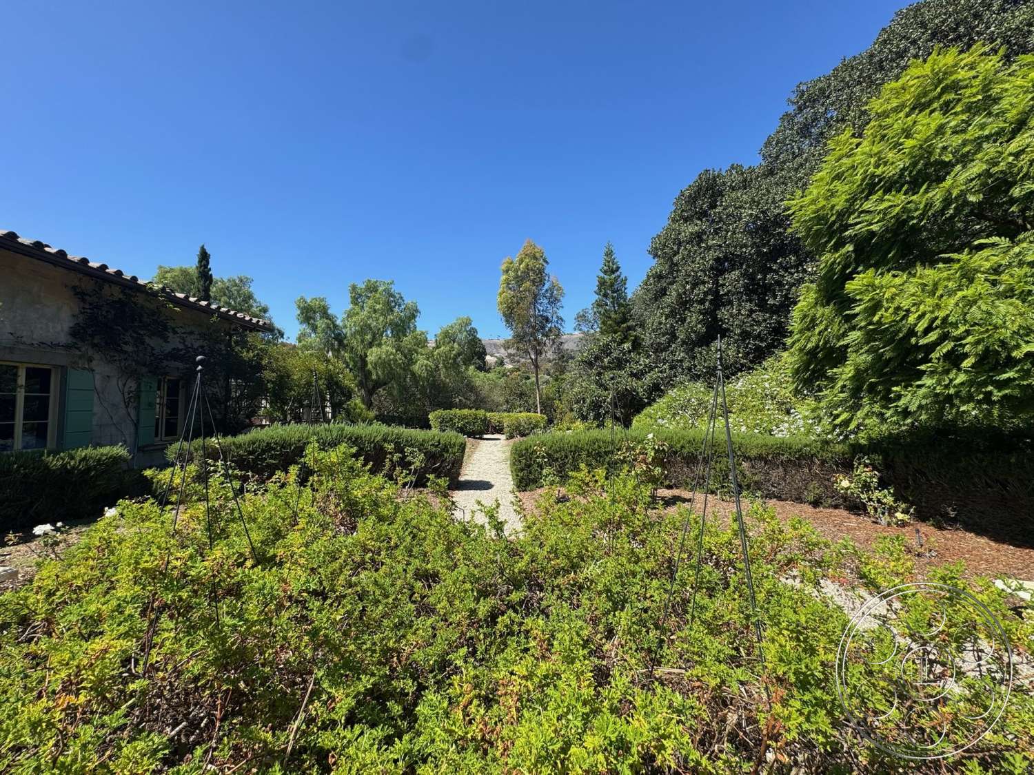 Farmstead Garden - exterior, gravel pathway, terracotta tile roof, stucco wall finish, landscaped garden beds, sunny outdoor environment, mediterranean architectural style, tree canopy overhead, pergola structure, drought-resistant planting, view of surrounding landscape, bush and shrub borders, cottage style home