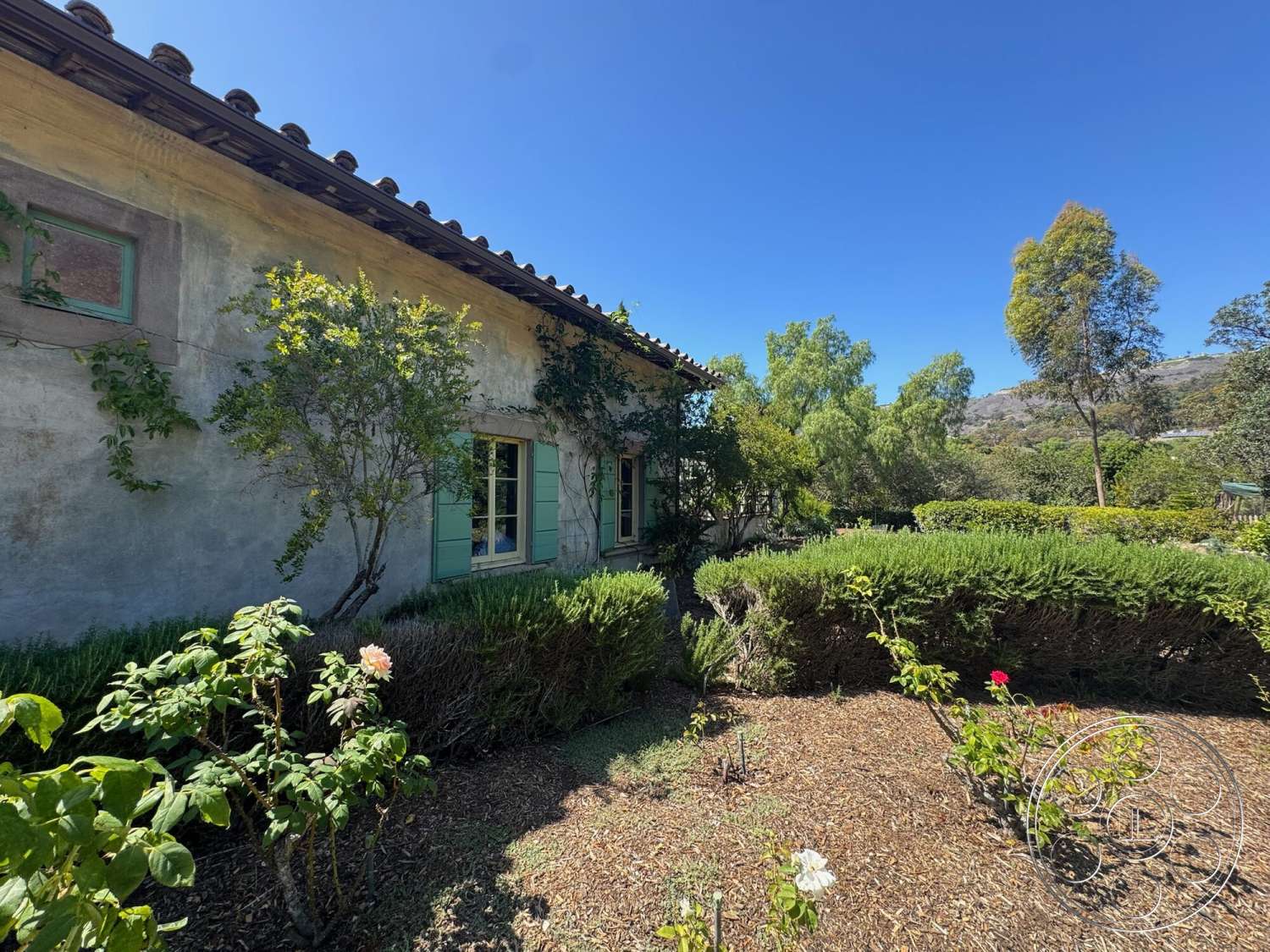 Farmstead Garden - exterior, landscaped garden, flowering shrubs, stucco wall finish, rustic architectural style, terra cotta tile roof, sunny hillside setting, hedge garden border, wood-framed windows with green shutters, Mediterranean home