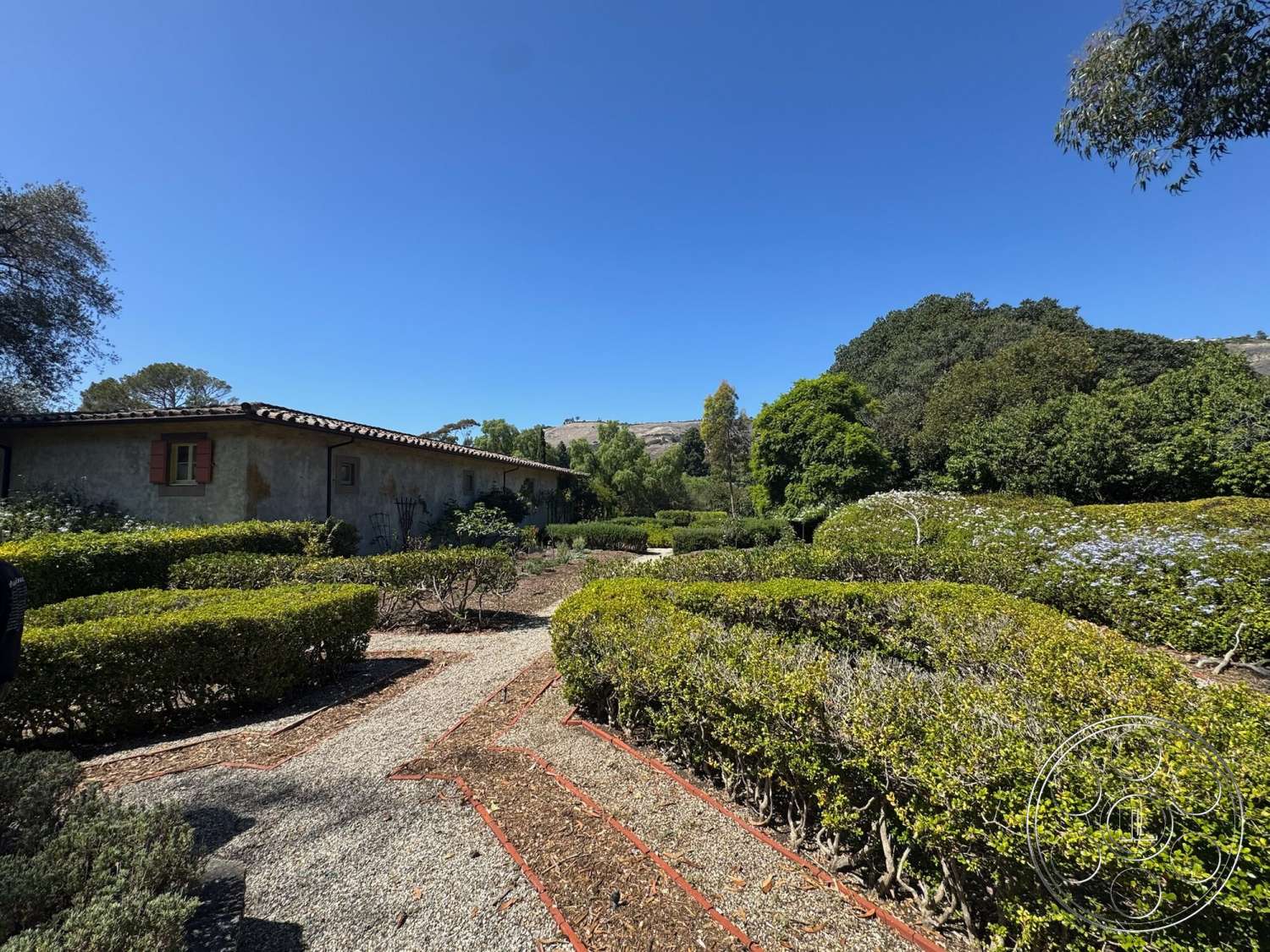 Farmstead Garden - exterior, gravel pathway, flowering shrubs, textured plaster walls, Mediterranean architectural style, clay tile roof, suburban garden, rolling hills background, red wooden shutters, trimmed hedge landscaping