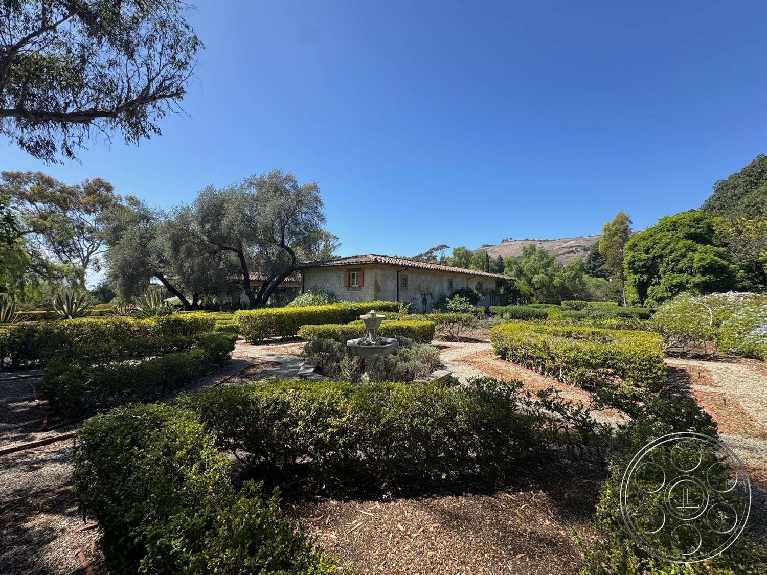 Farmstead Garden - exterior, lush greenery, outdoor seating area, surrounding hillside, gravel pathways, stucco walls, tile roof, arched windows, boxwood hedges, Mediterranean architectural style, circular fountain, dry landscaping, landscape gardens