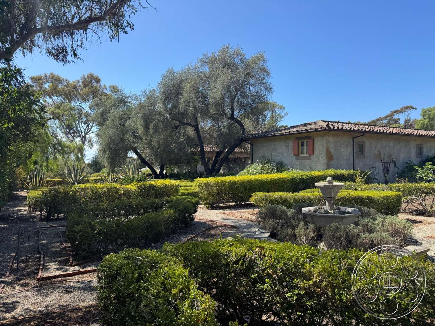 Farmstead Garden - exterior, landscaped garden, lush greenery, gravel pathways, stucco walls, desert plants, Mediterranean architectural style, clay tile roof, hedge borders, mature olive trees, horizontal window design, stone fountain feature, wood window shutters