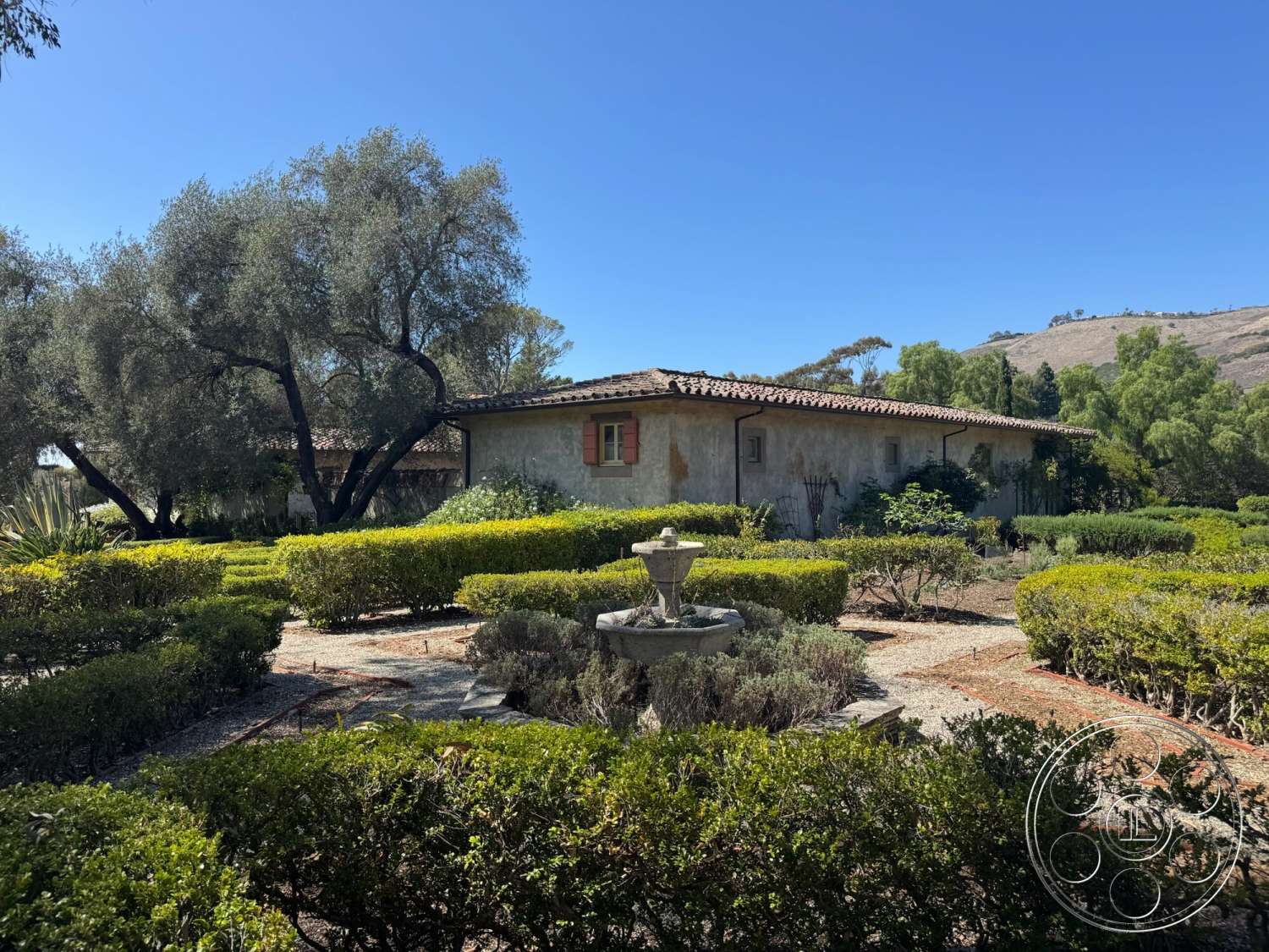 Farmstead Garden - exterior, landscaped garden, gravel pathway, mountain backdrop, red tile roof, olive trees, boxwood hedges, ornamental fountain feature, stucco house walls, terracotta window shutters