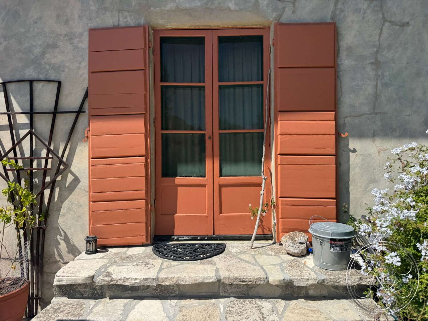 Farmstead Garden - exterior, concrete steps, plaster wall finish, stone patio flooring, flowering plants in pots, wooden door with glass panels, orange painted wooden shutters, decorative metal wall trellis, rustic metal bucket, black decorative doormat