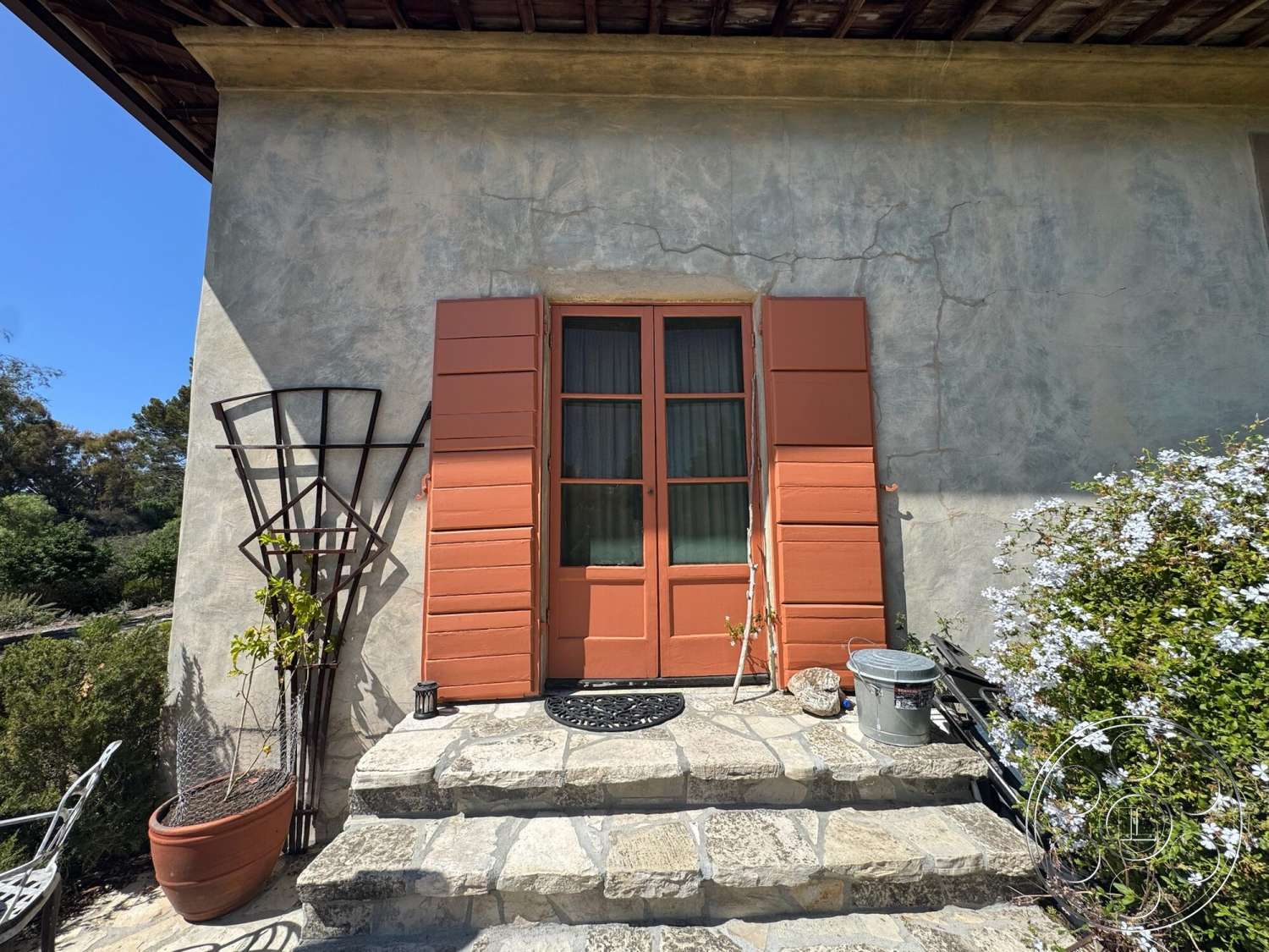 Farmstead Garden - exterior, flowering shrubs, flagstone patio, stucco wall finish, natural landscape setting, double door entrance, terracotta colored wooden shutters, stone step entryway, metal trellis with vine plant