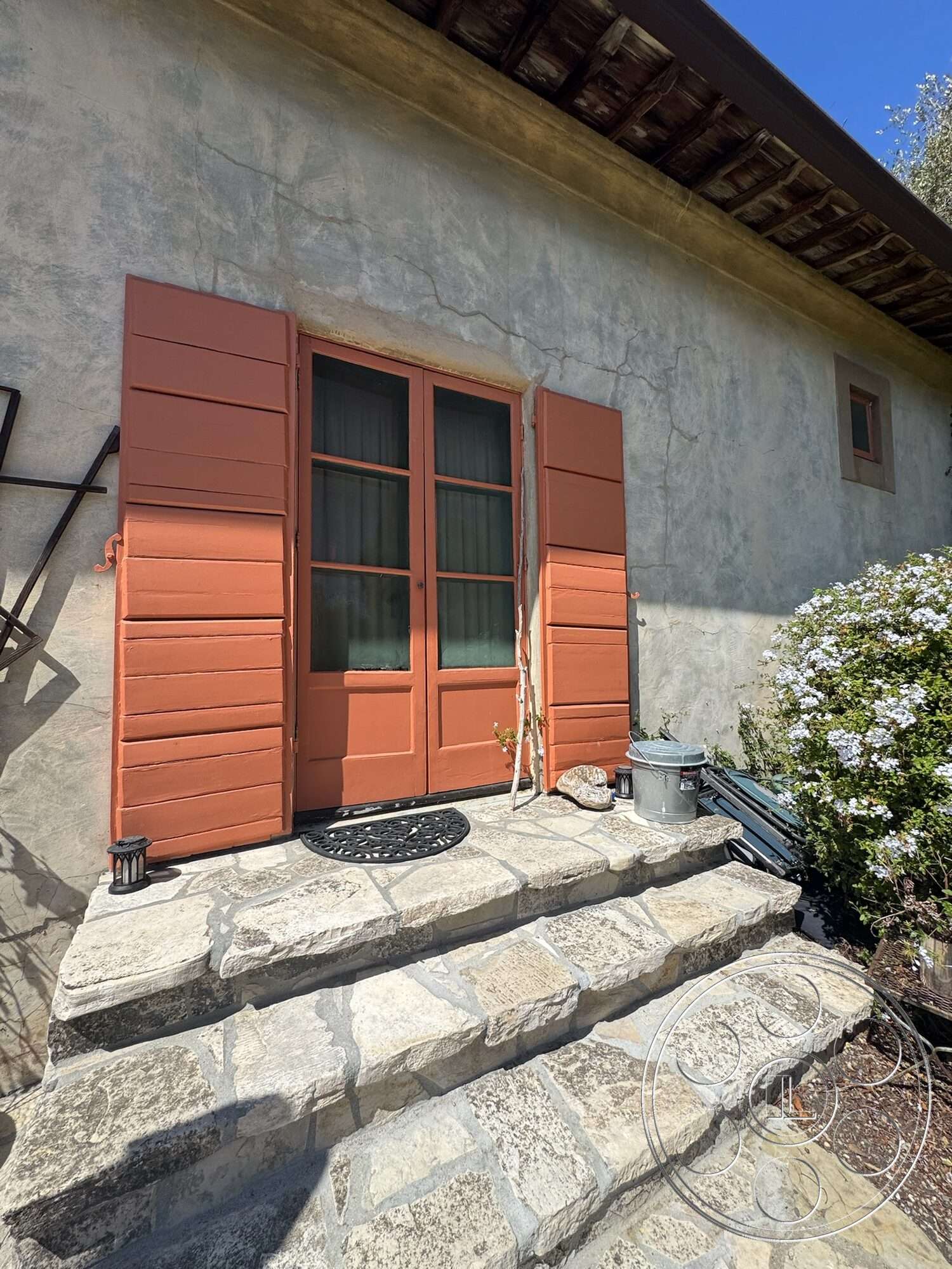Farmstead Garden - exterior, wooden ceiling beams, stone steps, double door entry, wooden window shutters, rustic architectural style, natural stone walkway, landscaped garden border, plastered wall finish, outdoor lantern lighting, orange painted window shutters, glass pane window