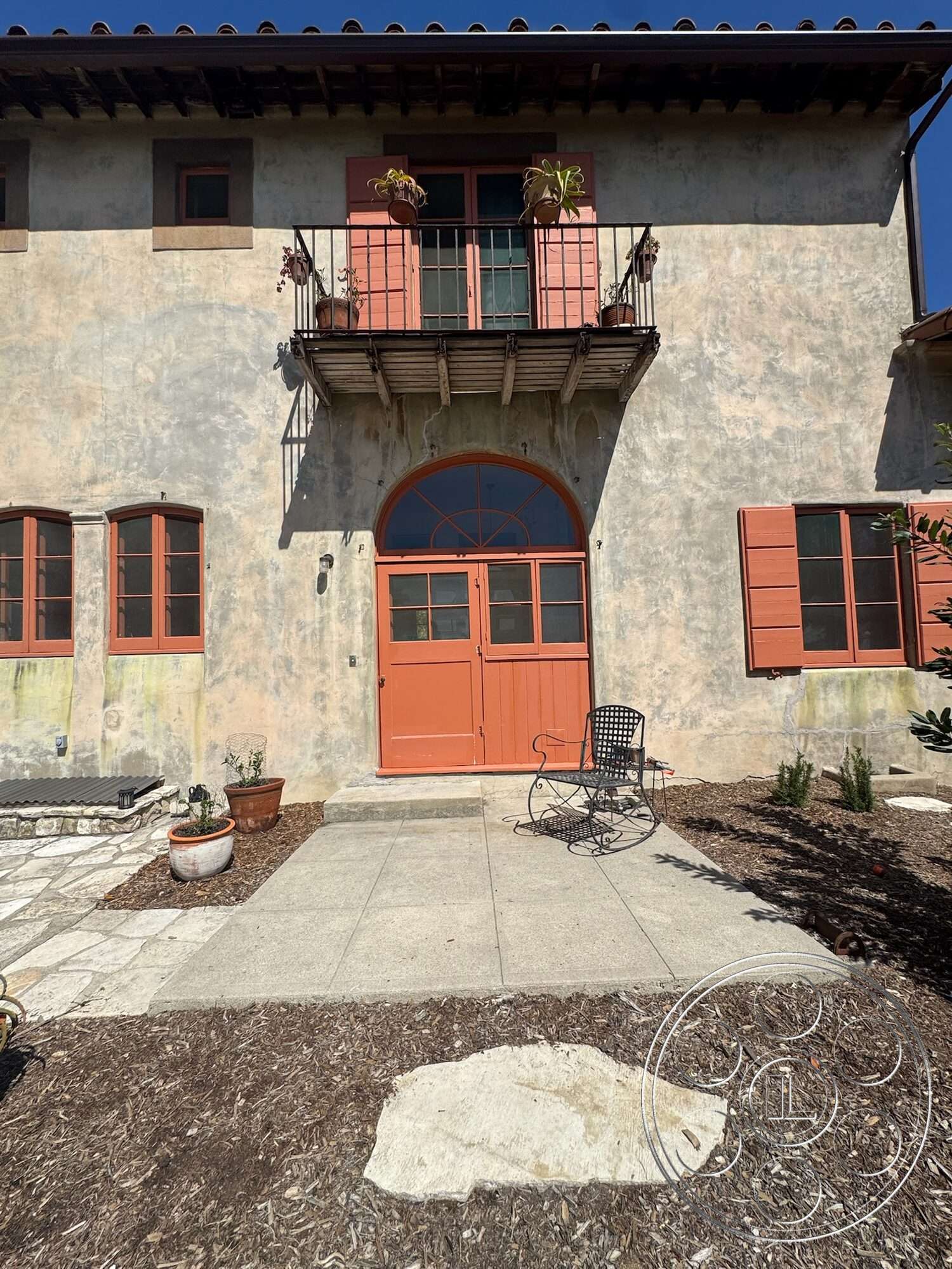 Farmstead Garden - exterior, exposed wooden beams, stone pathway, terracotta tile roof, Mediterranean architectural style, modern outdoor seating, stucco exterior walls, arched front door, rectangular concrete patio, wood balcony with railing, orange double doors, multicolored gravel landscaping, wood accents on windows, potted plants on balcony