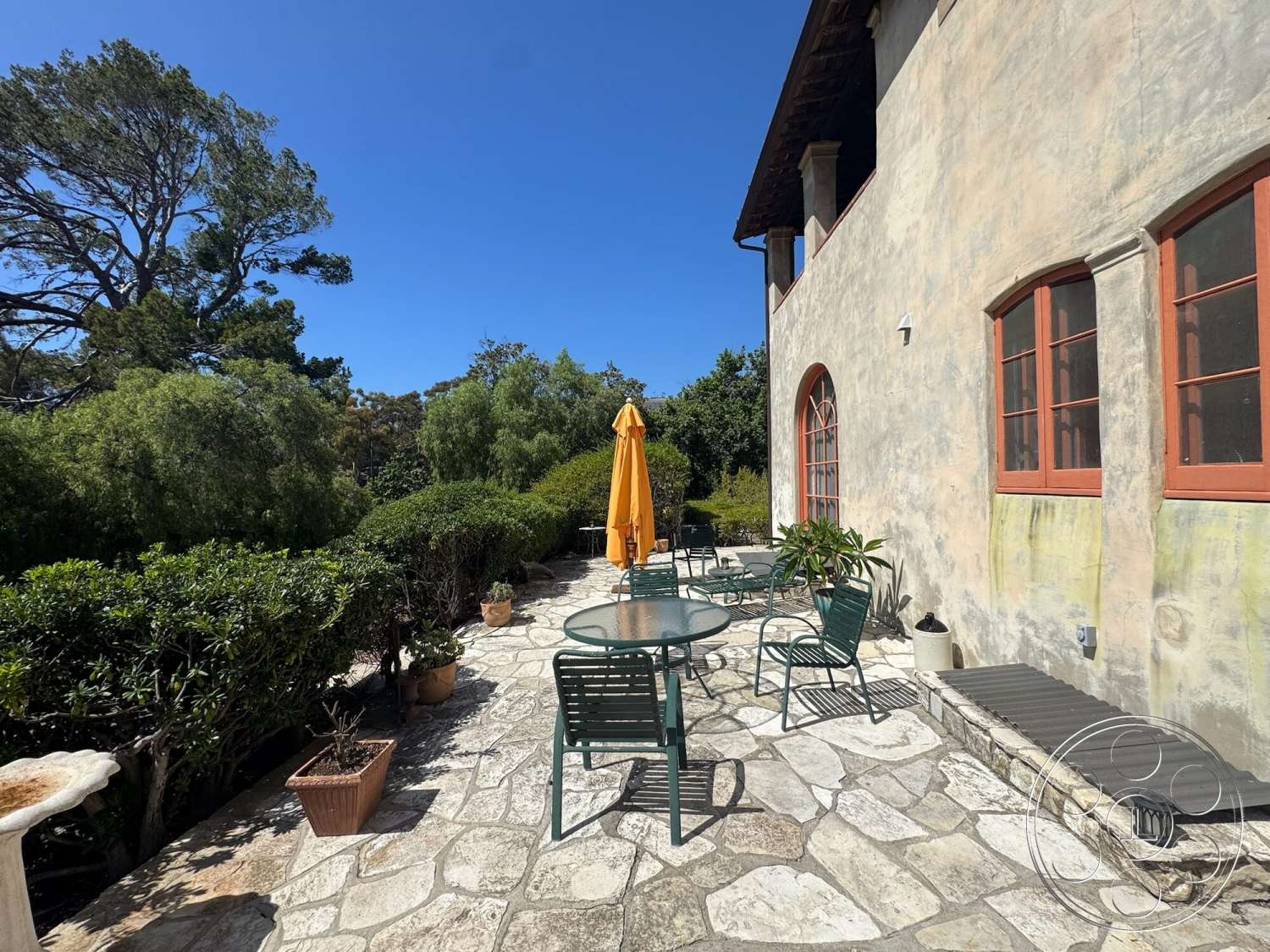 Farmstead Garden - exterior, potted plants, landscaped greenery, metal outdoor furniture, terracotta roof tiles, stucco walls, arched doorways, wooden window frames, sunny outdoor setting, outdoor umbrella, stone patio flooring, circular stone table