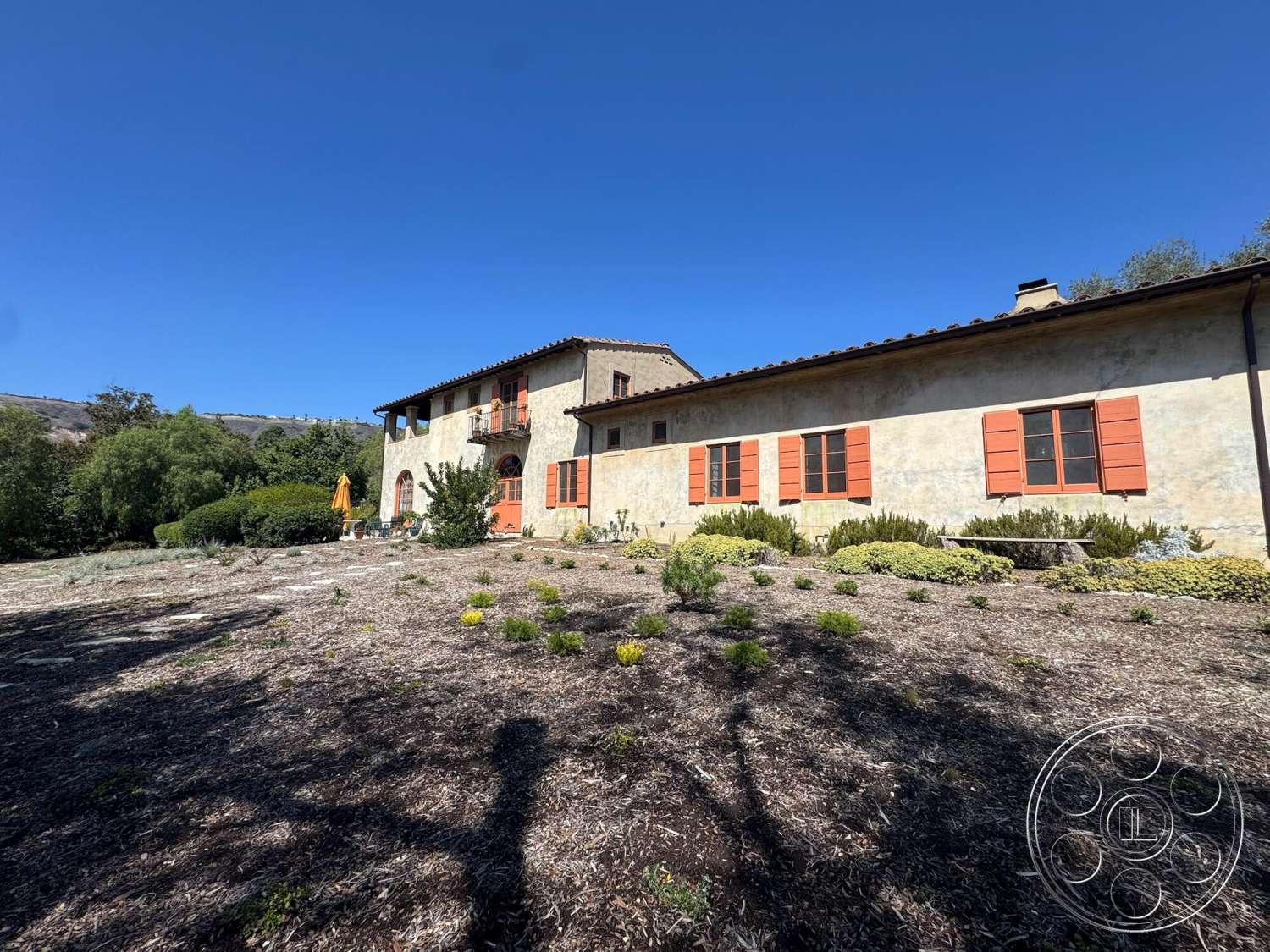 Farmstead Garden - exterior, landscaped front yard, terracotta roof tiles, Mediterranean architectural style, two-story home, stucco exterior walls, sunny hillside location, red wooden window shutters, stone landscaping borders, gravel garden areas, vertical balcony railing, open garden patio