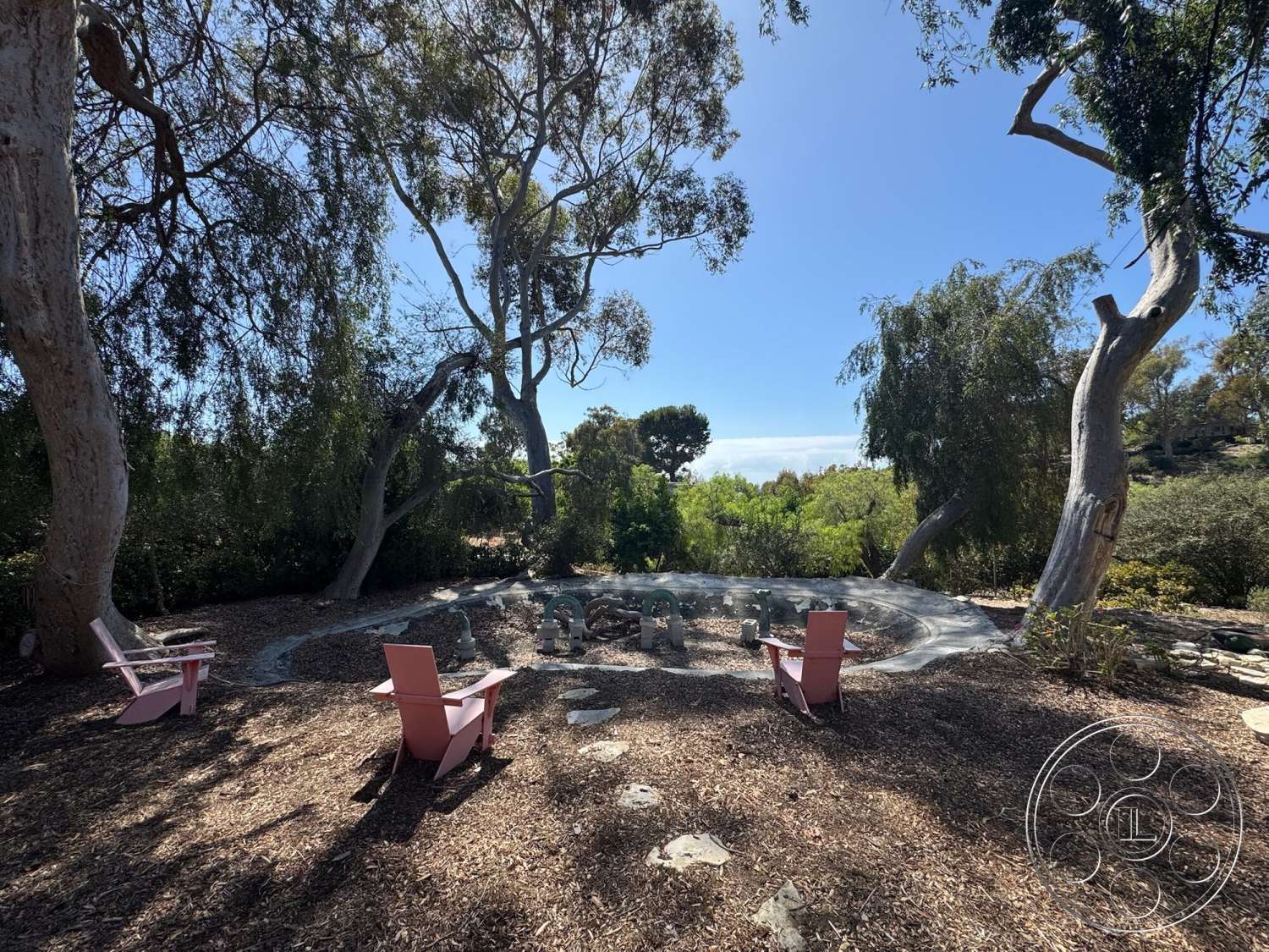 Farmstead Garden - exterior, landscaped garden, lush greenery backdrop, eucalyptus trees, pink adirondack chairs, sunny outdoor setting, wooden patio flooring, open sky view, natural stone fire pit, minimalist outdoor seating area, round fire pit design, wood chip mulch ground cover, relaxed gathering space