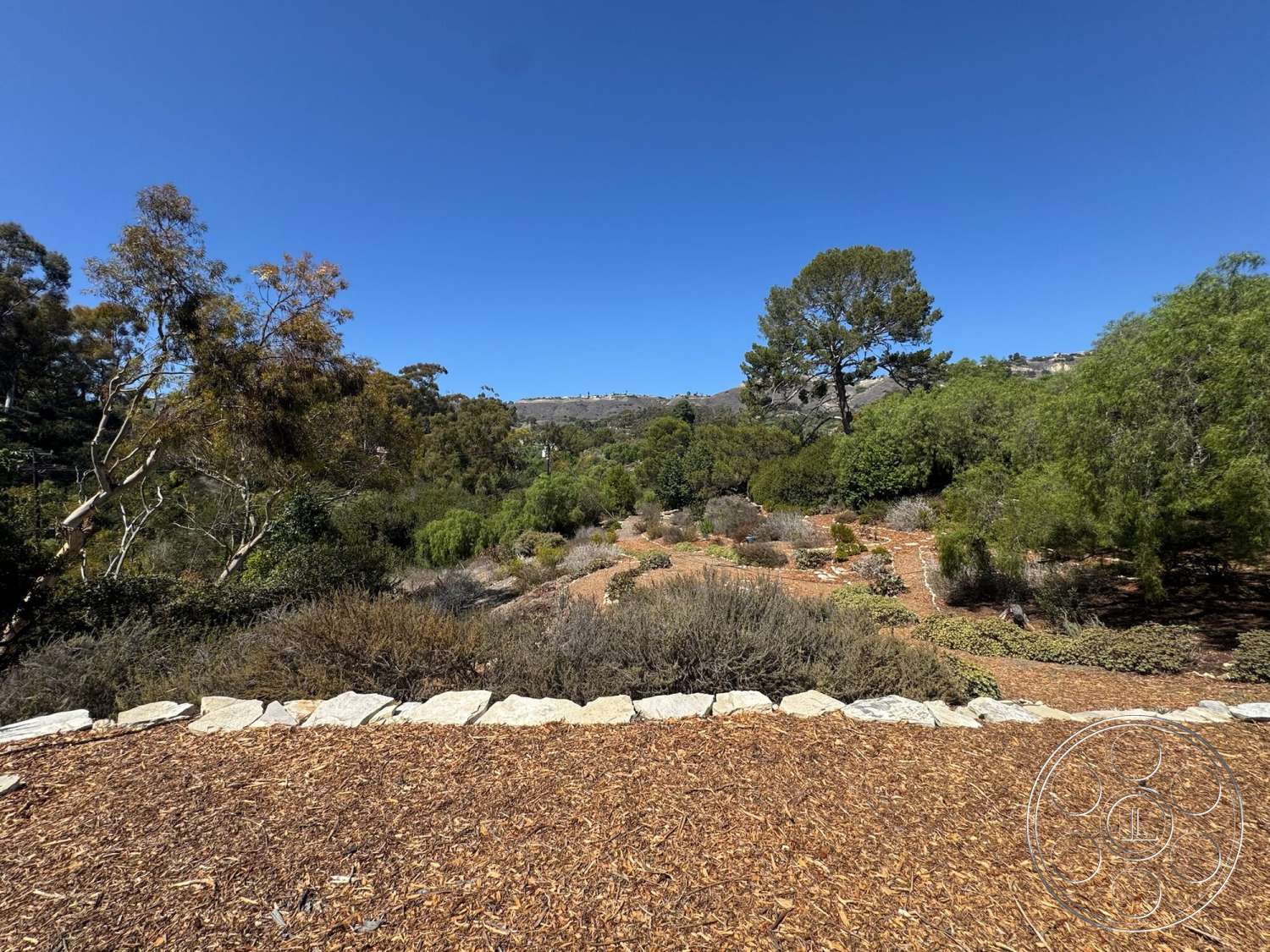 Farmstead Garden - exterior, clear blue sky, mixed vegetation, mulch ground cover, oak trees, scenic hillside view, low-maintenance landscape design, natural stone wall border, native shrub landscaping, meandering pathways