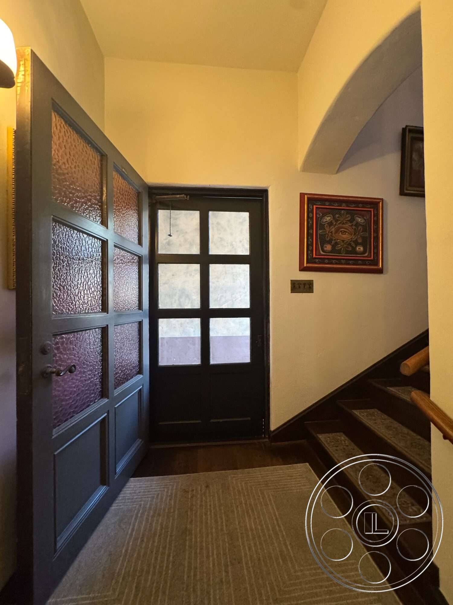 Farmstead Garden - neutral wall color, framed wall art, textured plaster walls, glass panel door, wooden front door, area rug with geometric pattern, dark wood staircase, arched doorframe, brushed metal wall switch