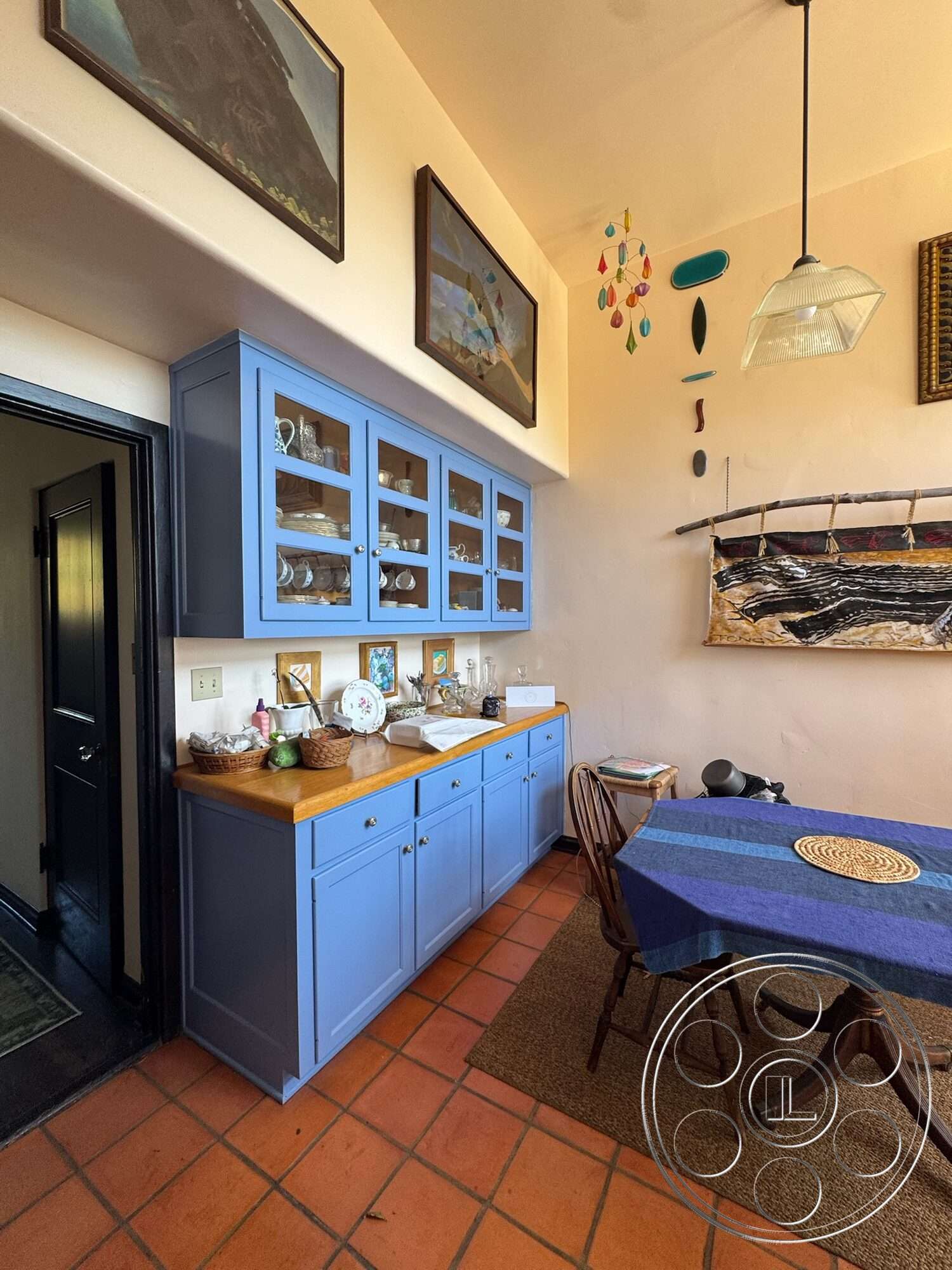 Farmstead Garden - open shelving, textured wall finish, high ceiling, black door, round dining table, hanging light fixture, wood countertop, framed wall art, colorful wall accents, decorative wall hanging, clay tile flooring, blue cabinetry kitchen cabinets, woven placemat, rustic dining chairs