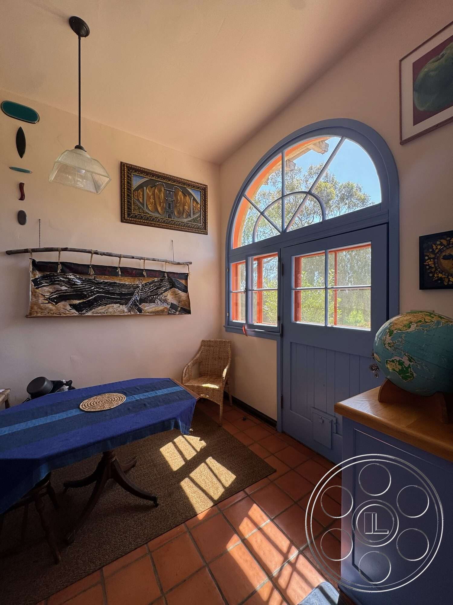 Farmstead Garden - natural light, woven chair, wooden dining table, hanging pendant light, casual dining area, colorful artwork, textured plaster walls, arched window, woven wall hanging, stool, blue accent wall, teracotta tile flooring, blue painted door, hexagonal globe