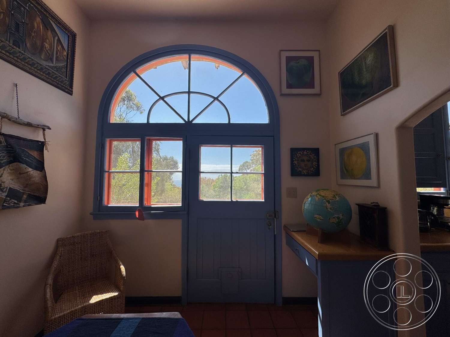 Farmstead Garden - woven chair, wooden countertop, white plaster walls, decorative globe, natural light from large window, framed artwork on walls, arched window with divided panes, blue painted wooden door, light terracotta tile flooring